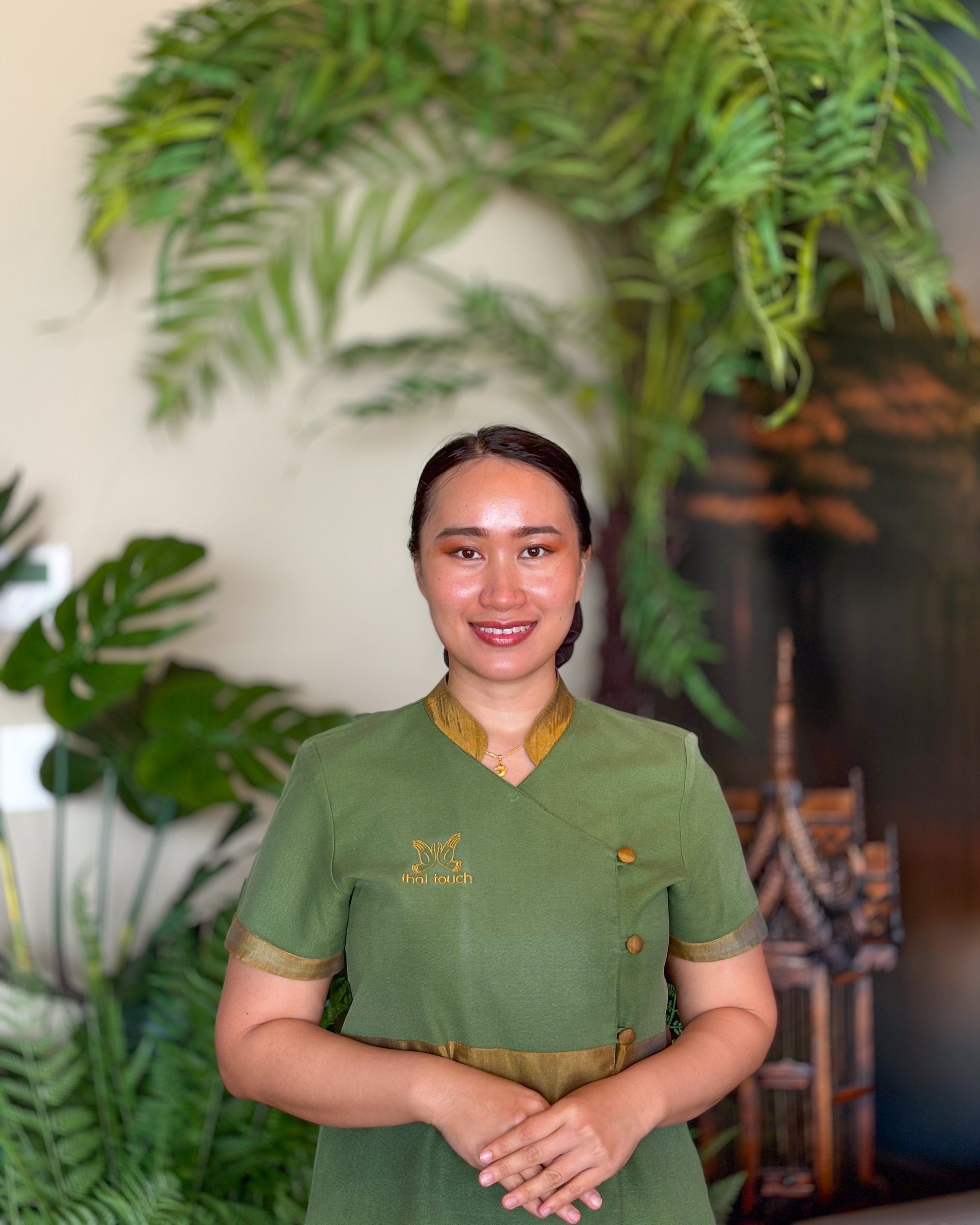 Thai Touch Massage Therapist, Anchy, standing indoors with large green plants and wooden furniture in the background.