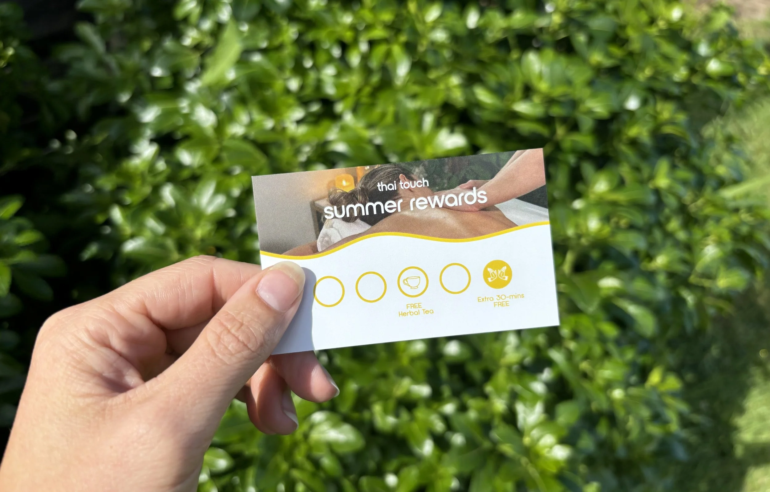 Hand holding a reward card for Thai Touch Summer Rewards, featuring a massage scene and icons for free herbal tea and extra 30-minute free session, in front of green foliage.
