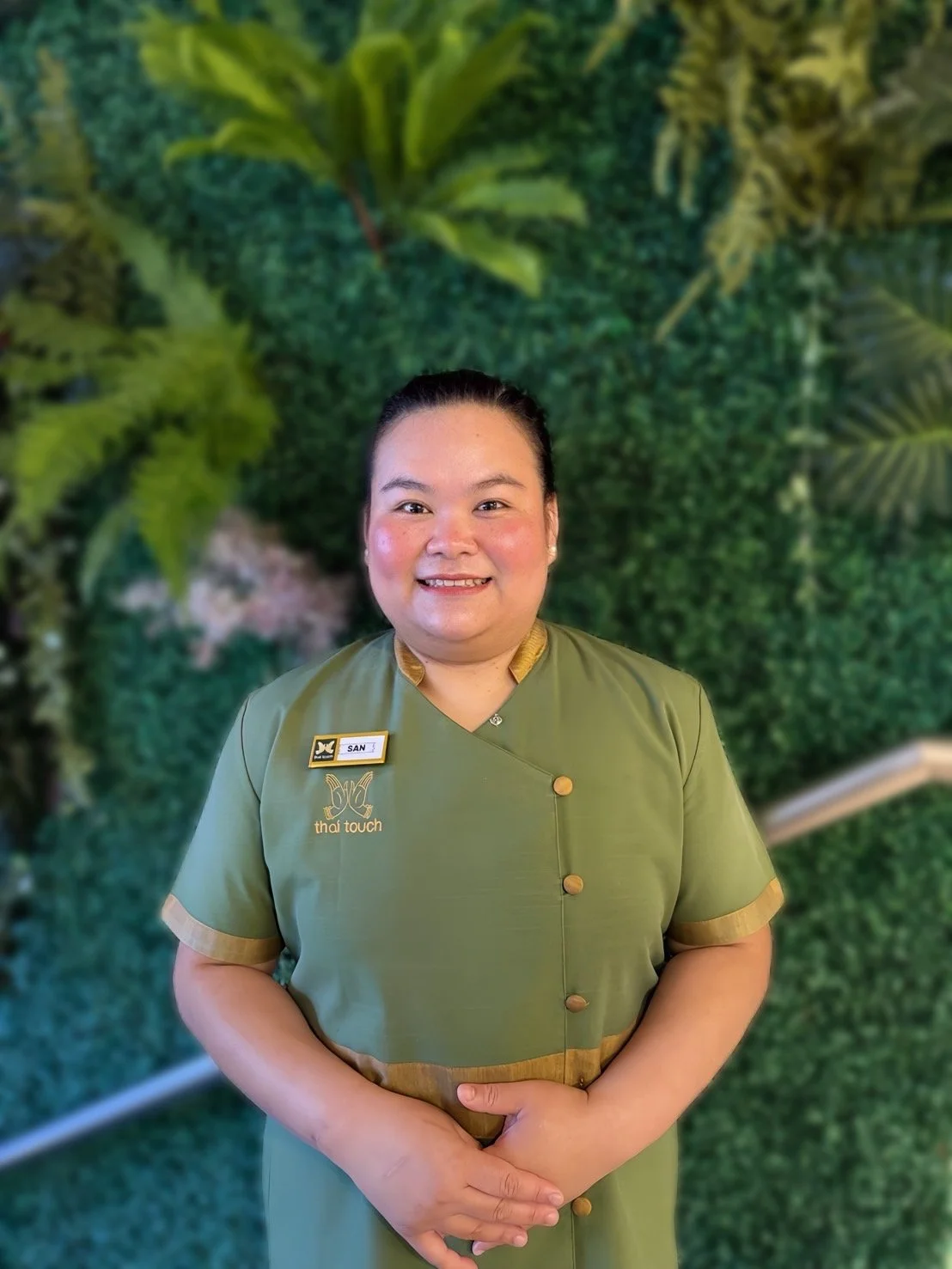 Thai Touch's San, standing in front of a lush green foliage background, smiling at the camera.