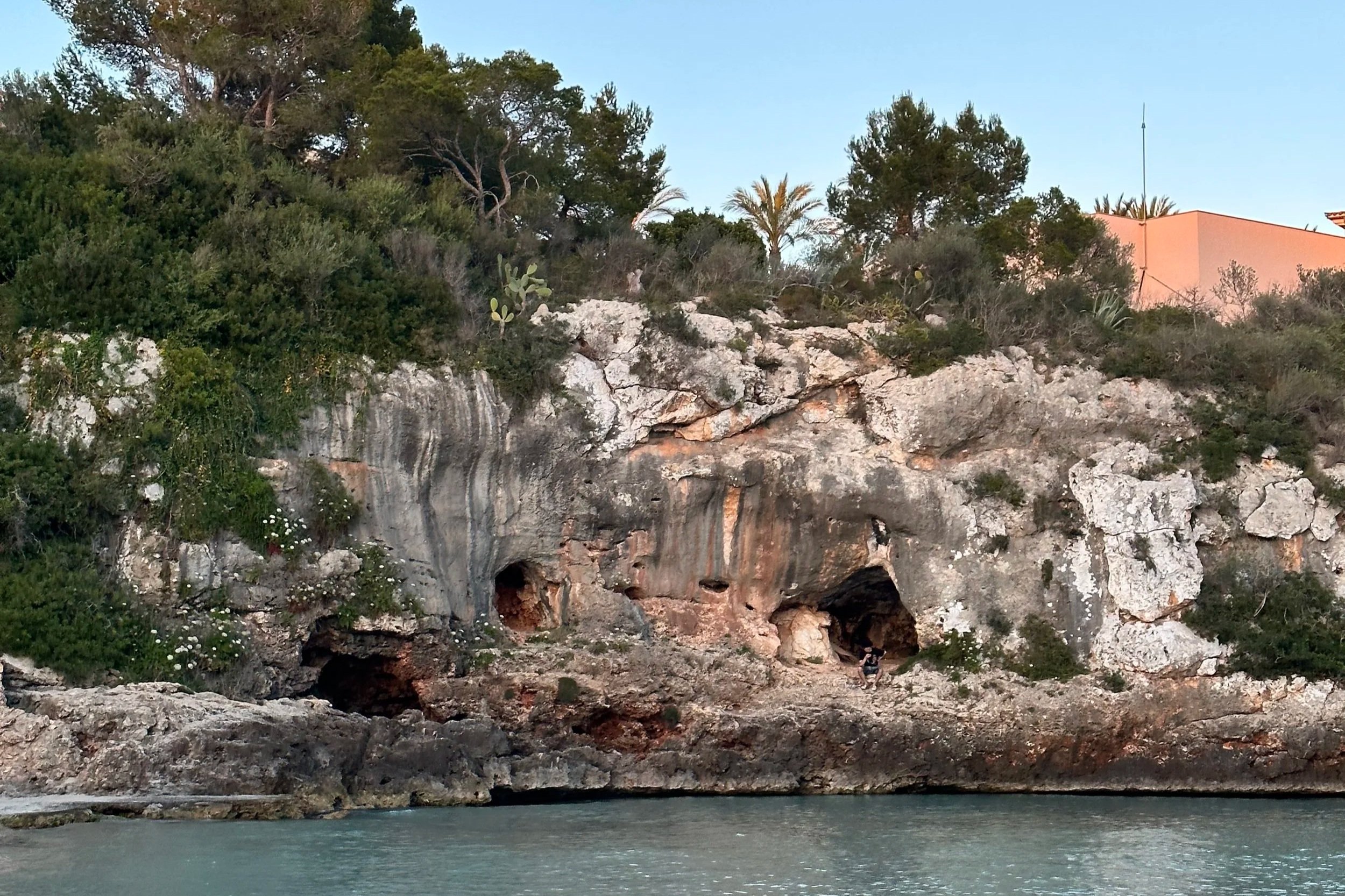 Rocky cliffside with caves above water, surrounded by green bushes and trees, with buildings in the background and a clear blue sky.