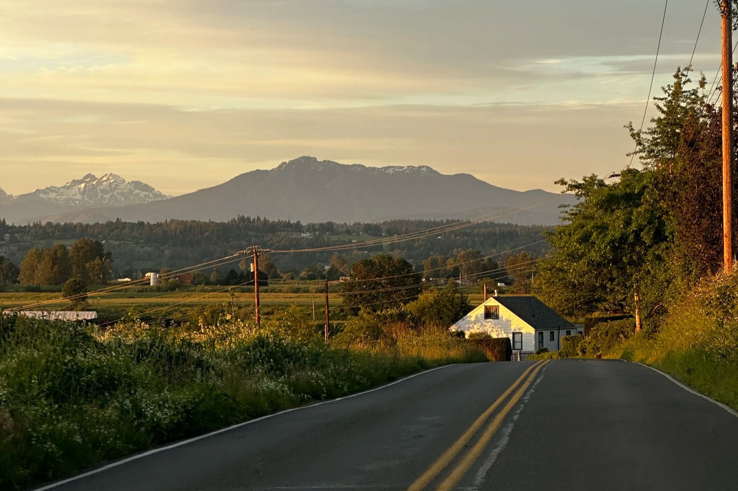 A rural road leading towards a white house, with green trees and fields on either side. In the background, there are mountains with snow on their peaks under a partly cloudy sky.