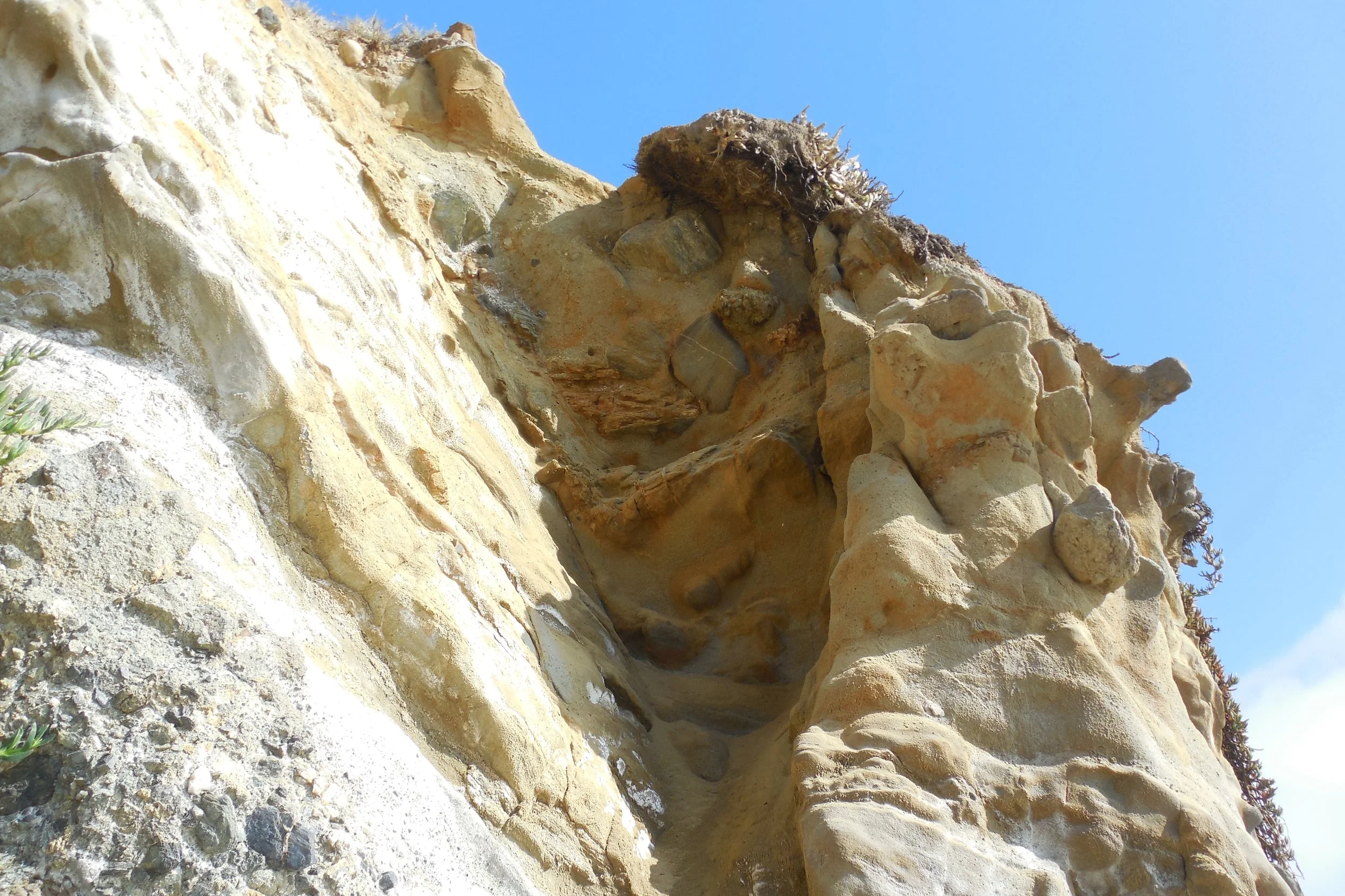 Close-up of a sandstone cliff with various textures and small rocks, against a clear blue sky.
