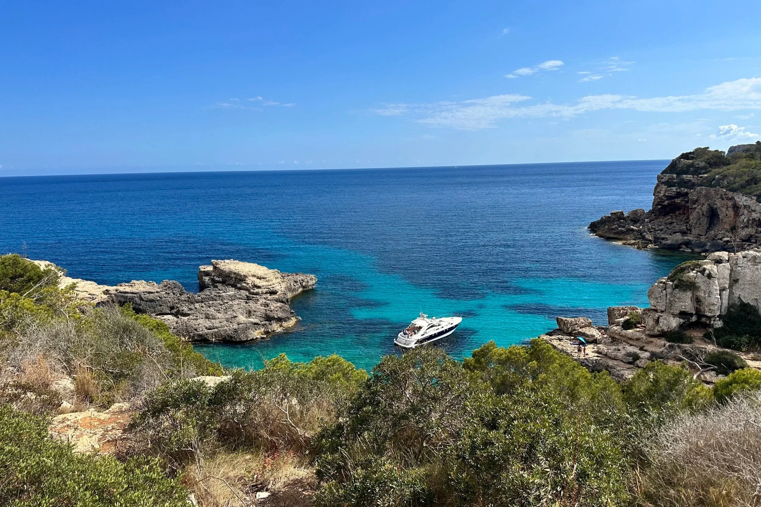 A scenic coastal view with clear blue waters, rocky cliffs, lush green vegetation, and a white yacht anchored near the shore.