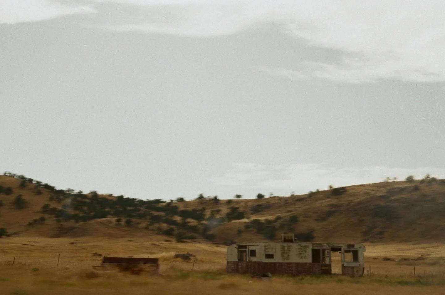 An abandoned trailer in a dry, hilly landscape with sparse trees and a cloudy sky.