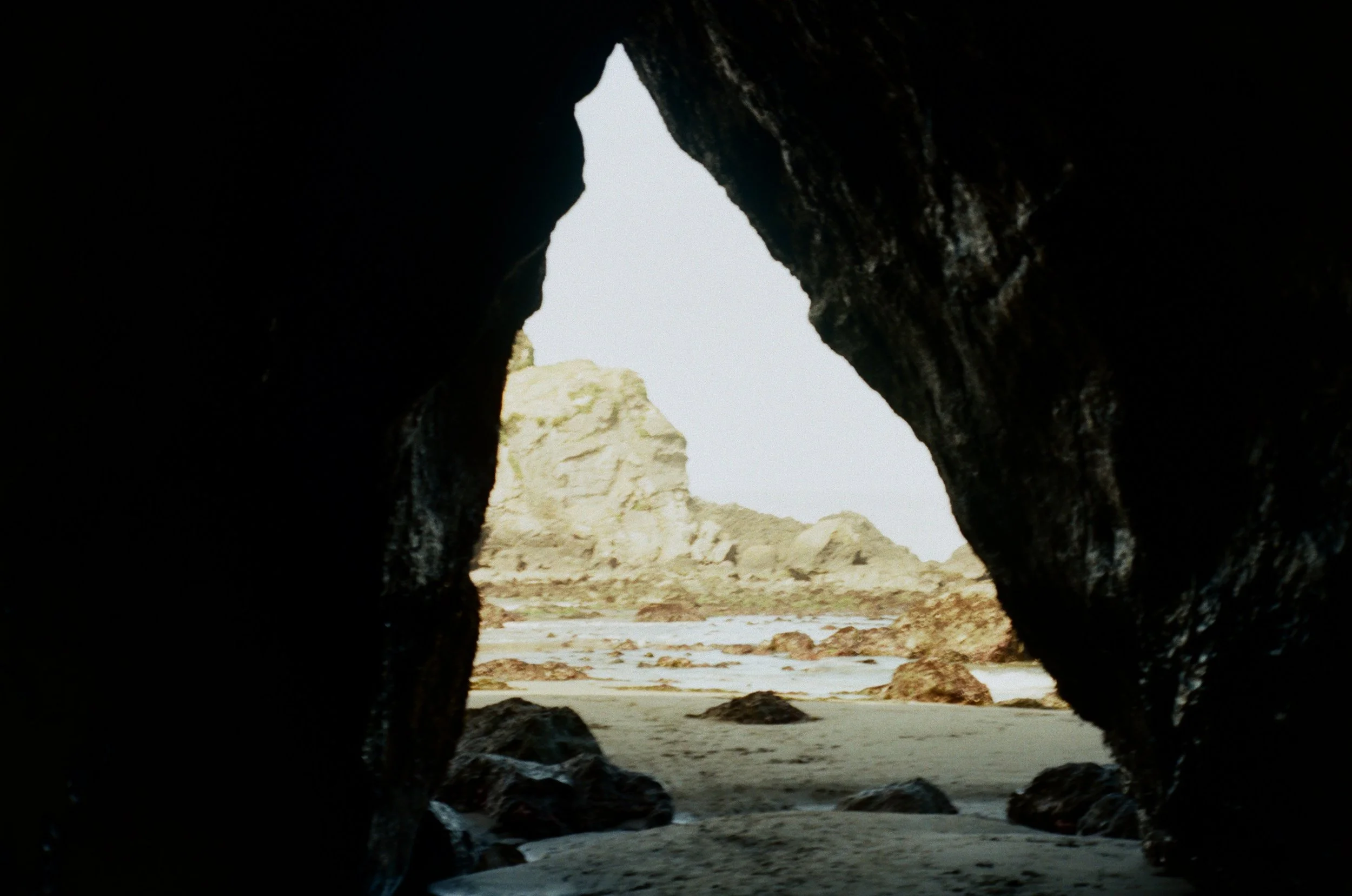View of a rocky coastal landscape seen through a natural rock arch or cave opening, with sandy beach and cliffs in the background.