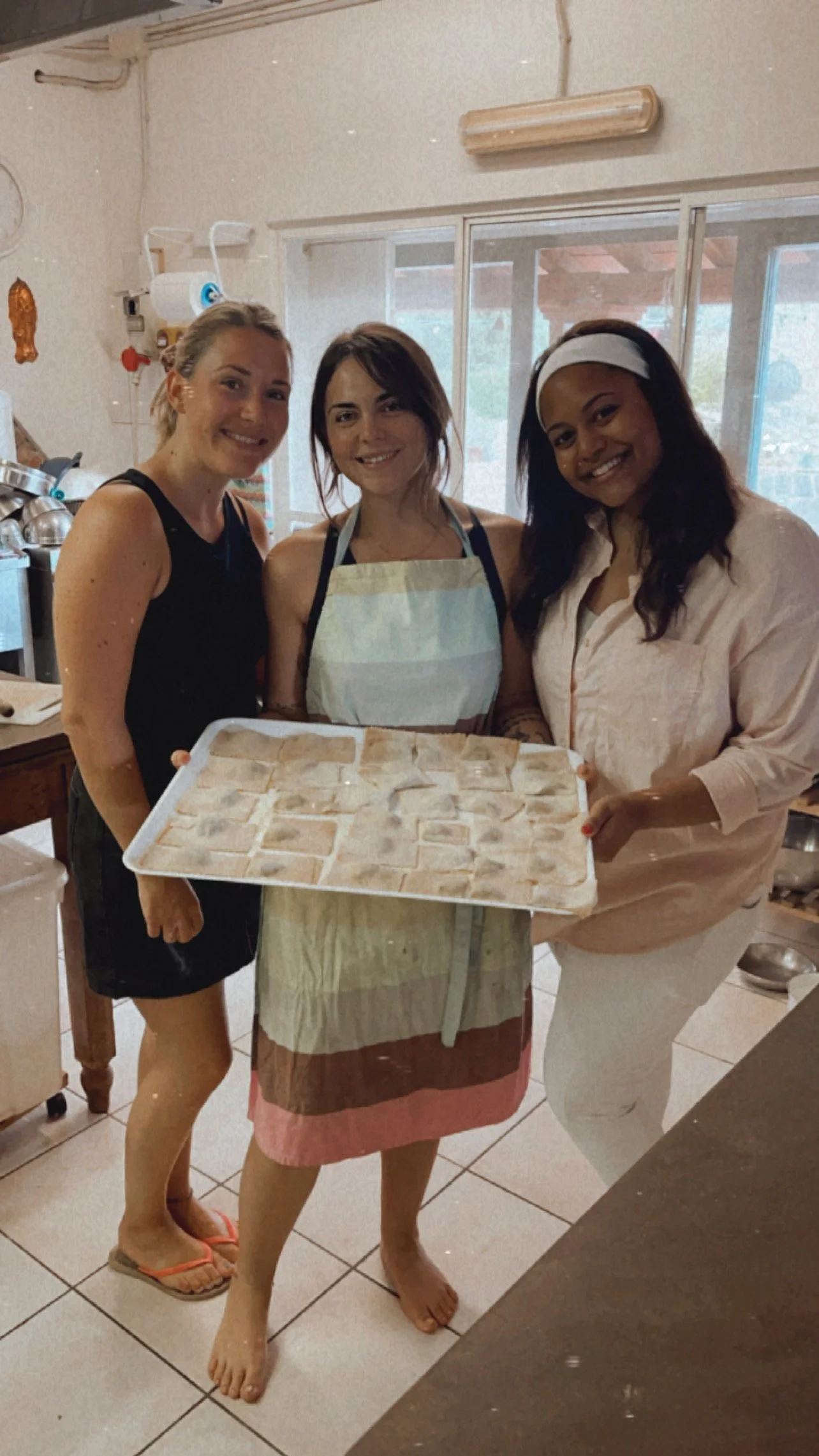 Three women standing in a kitchen, holding a baking tray with dough-cut cookies. One woman is wearing an apron, and they are smiling for the photo.