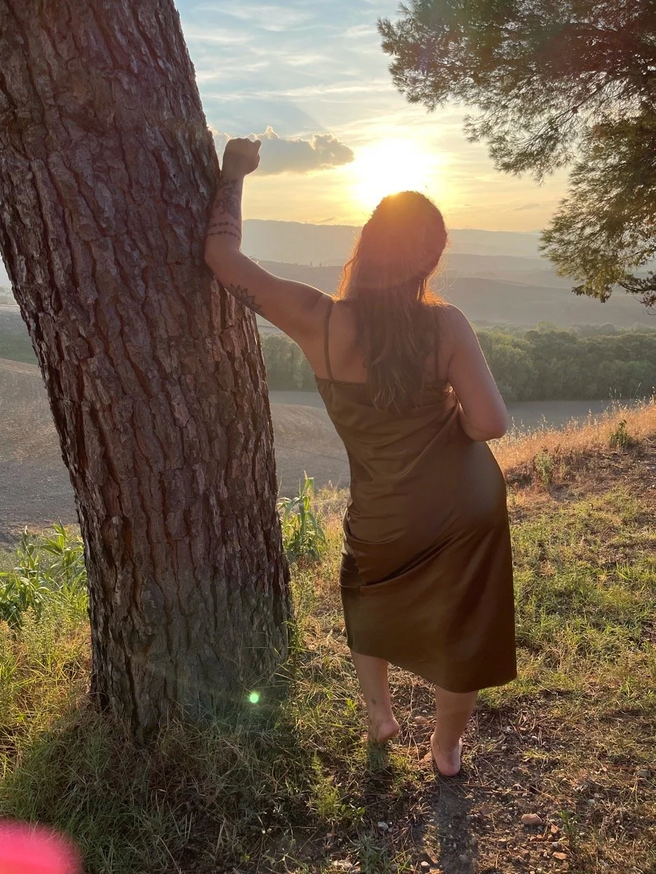 A woman in a brown dress with tattoos on her arm stands barefoot on a grassy hill, leaning against a tree at sunset, overlooking a landscape of trees and rolling hills.