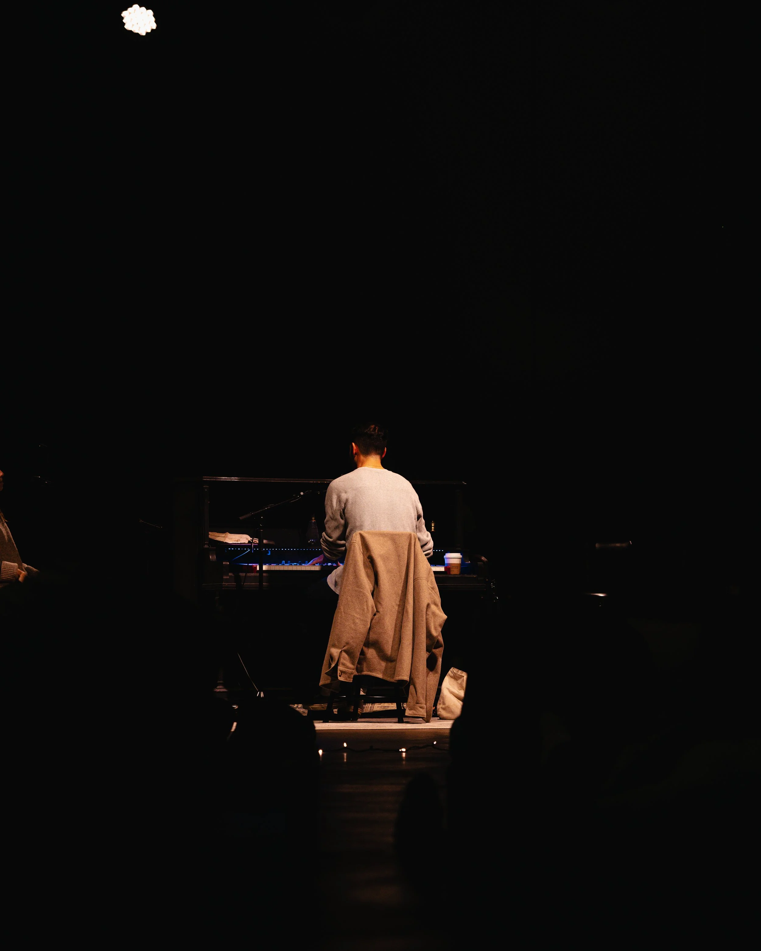Person playing piano on stage, back turned, with a beige coat draped over chair, dimly lit performance setting.