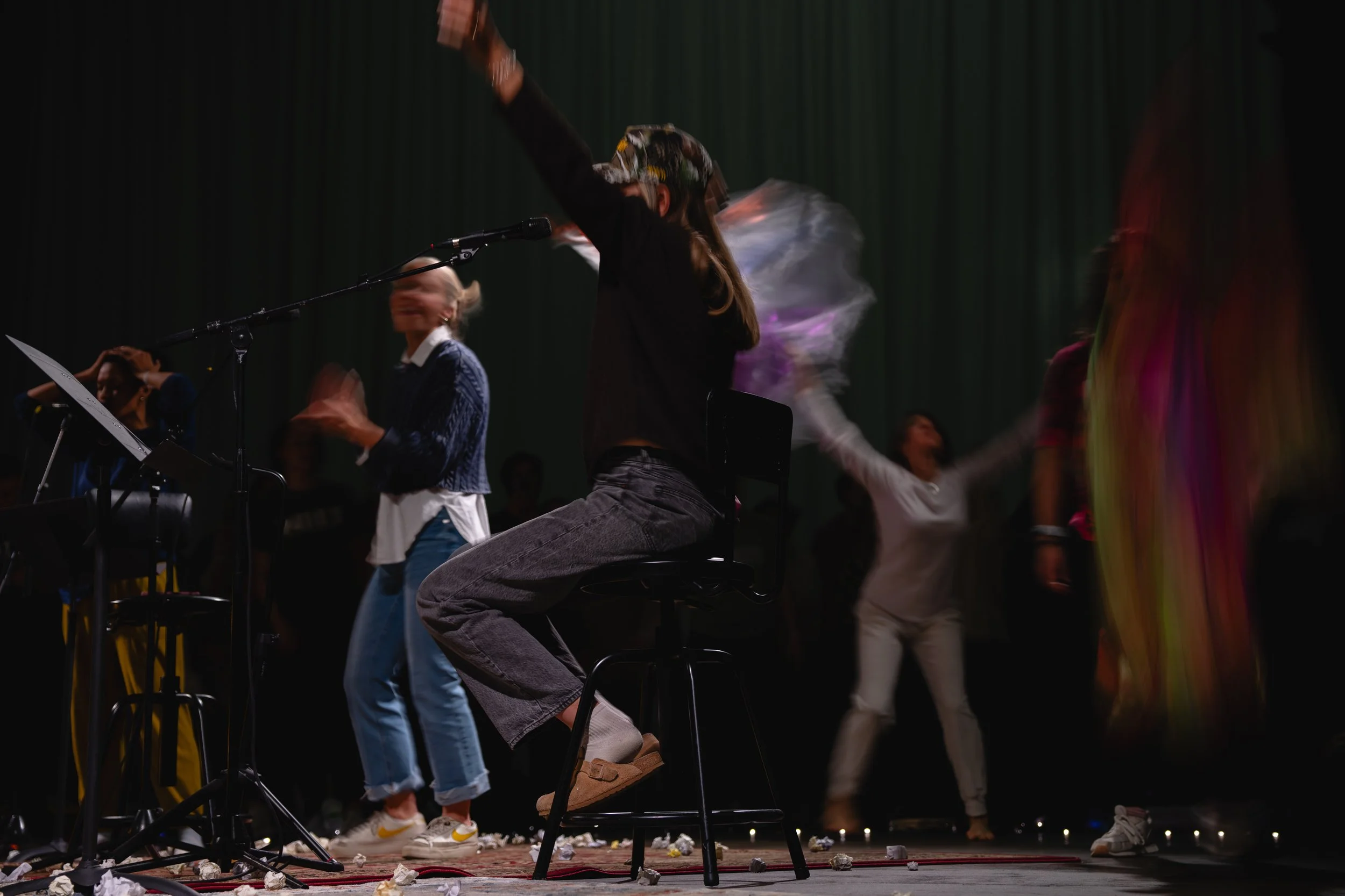 Group of people dancing and sitting on stage with music equipment, some holding fabric or scarves, with a dark curtain background and stage lighting.