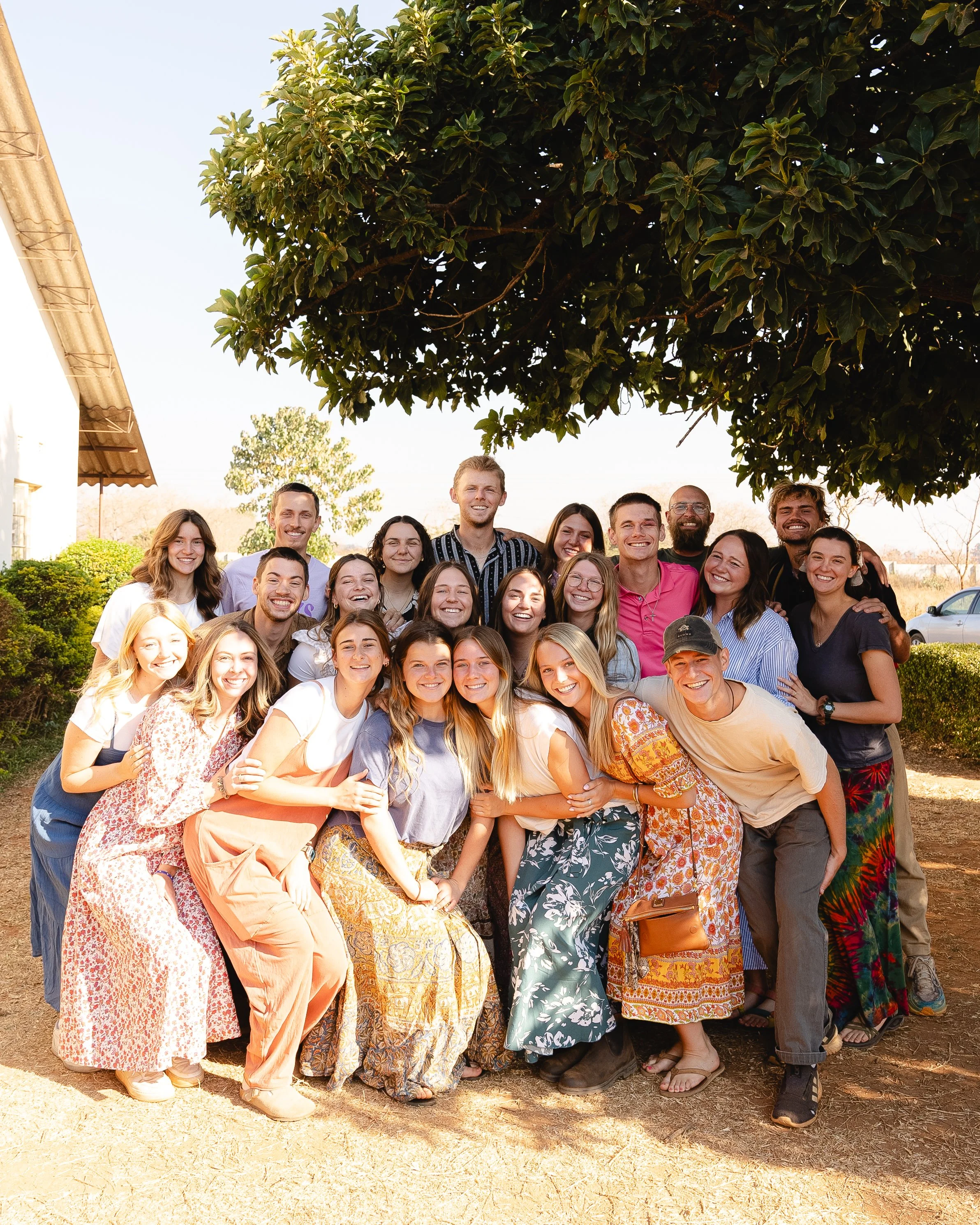 A group of smiling young adults gathered outdoors under a large leafy tree, posing for a photo with some people crouching in front and others standing behind.