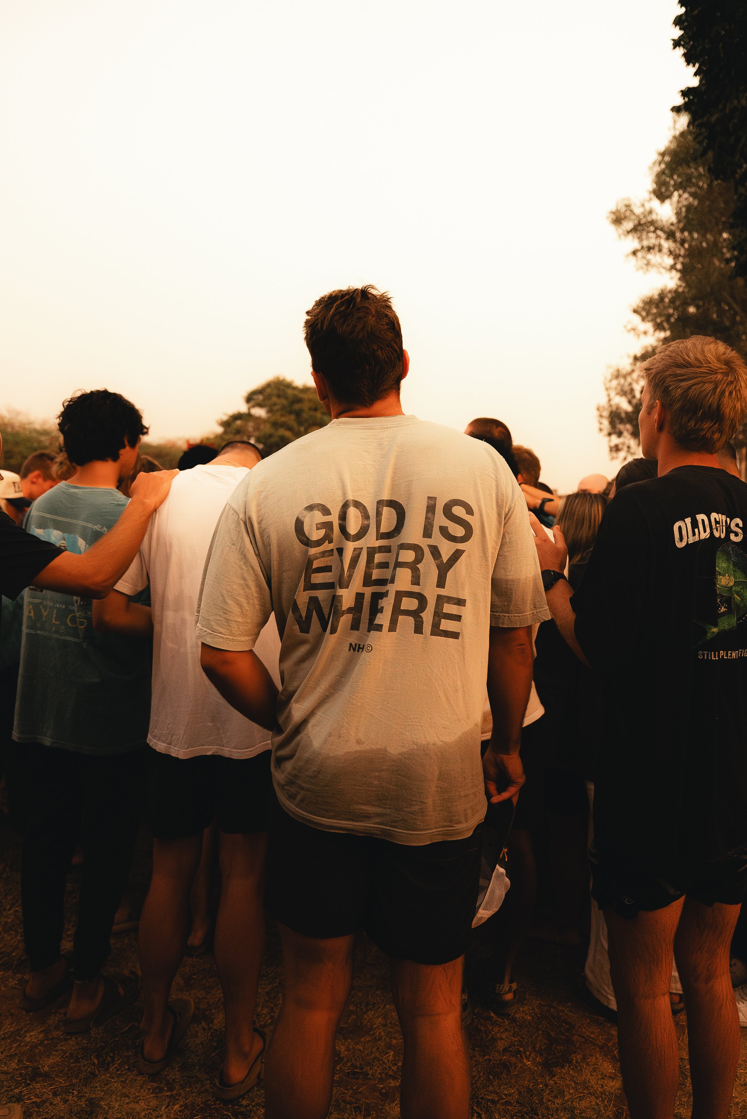 A group of people gathered outdoors during sunset, with one person in the center wearing a T-shirt that reads 'GOD IS EVERY WHERE' on the back.