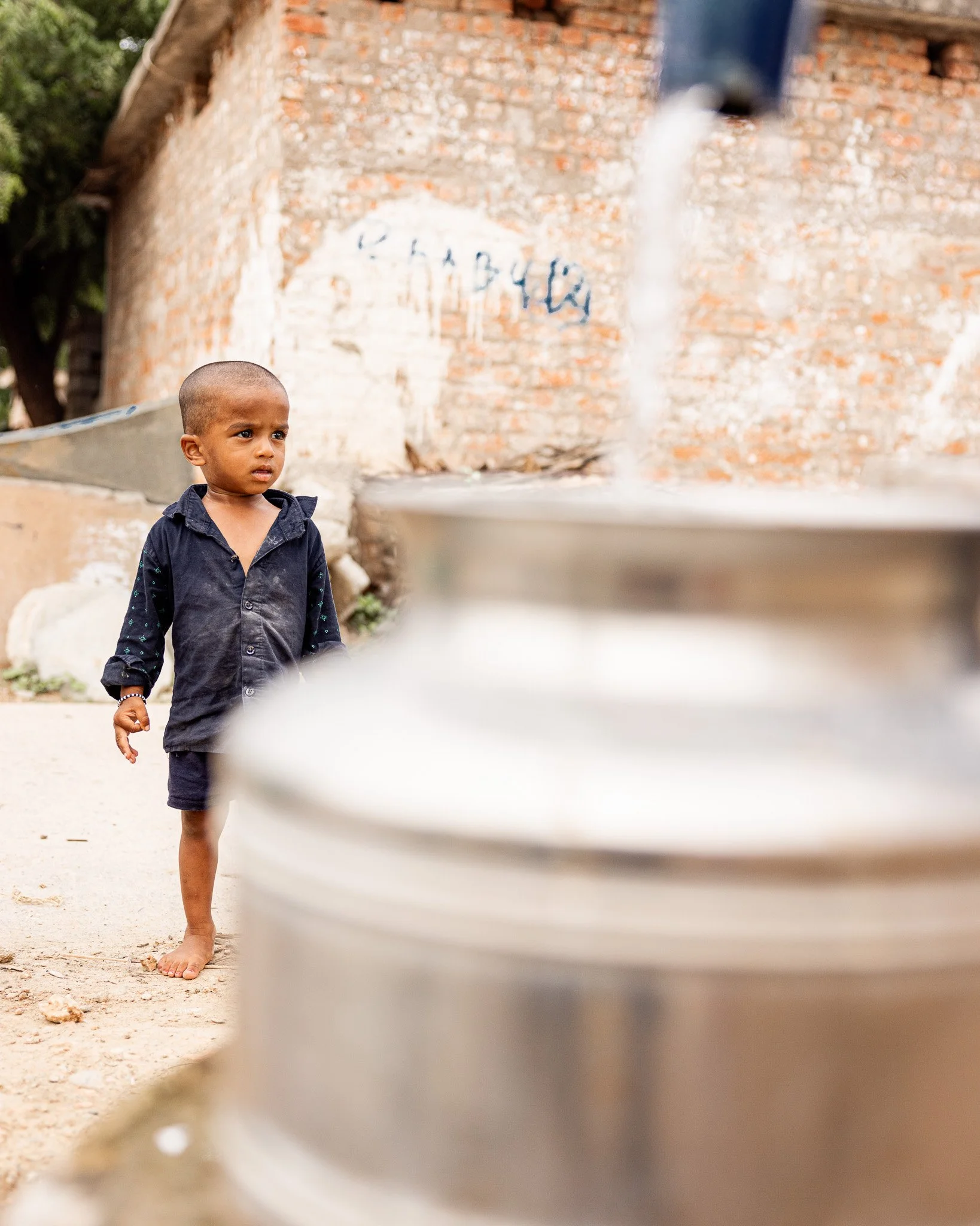 A young boy with a shaved head, wearing a black shirt and shorts, stands barefoot on a dirt ground and looks at a water spout or pipe. There are large metal containers in the foreground, and a brick wall with faded writing in the background.