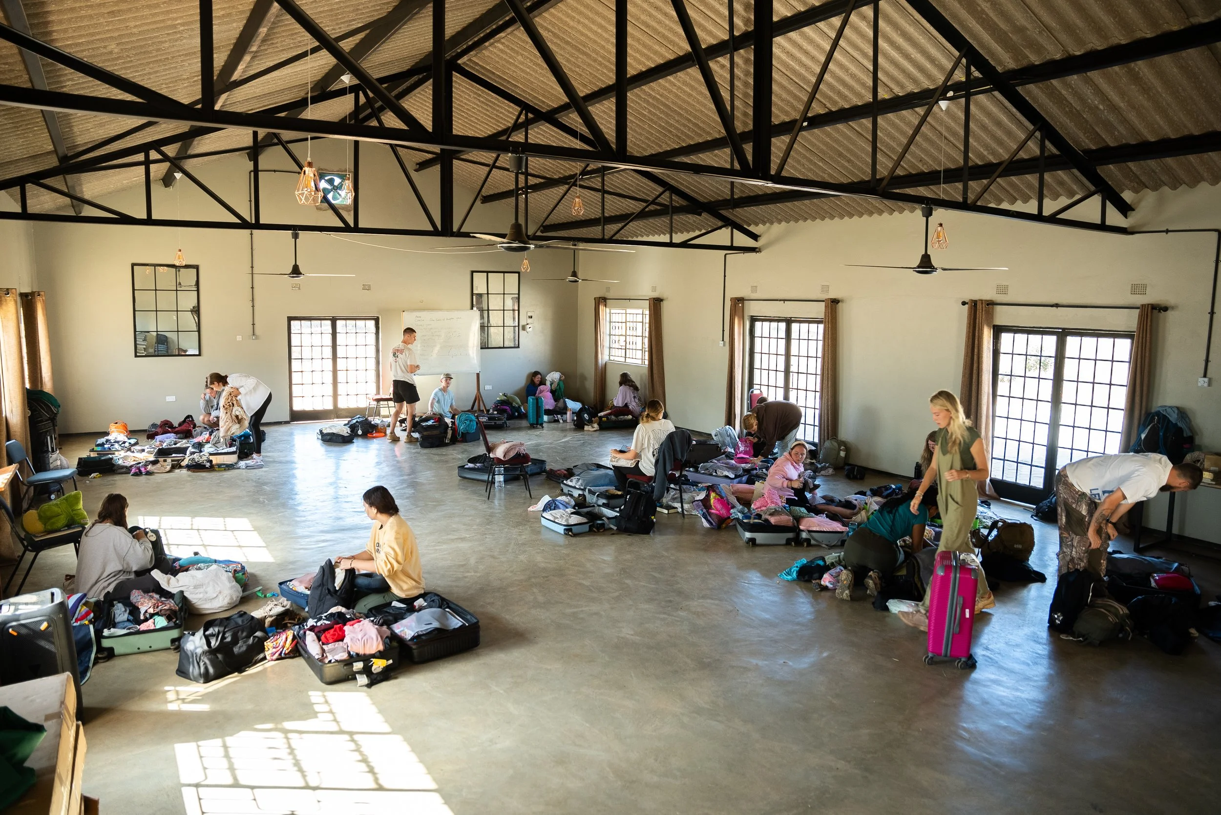 People inside a spacious, well-lit room with high ceilings, large windows, and a tiled floor, packing or unpacking suitcases and bags.
