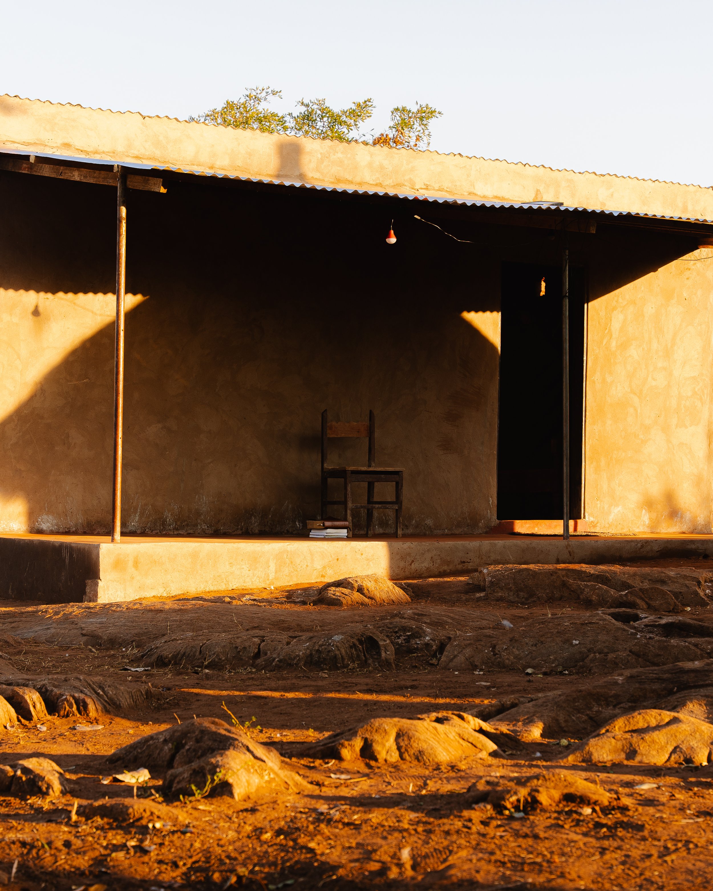 A simple, unfinished house with a corrugated metal roof, a black door, a wooden chair, and a small pile of books near the chair, set against a dirt ground with rocks, bathed in warm sunlight.