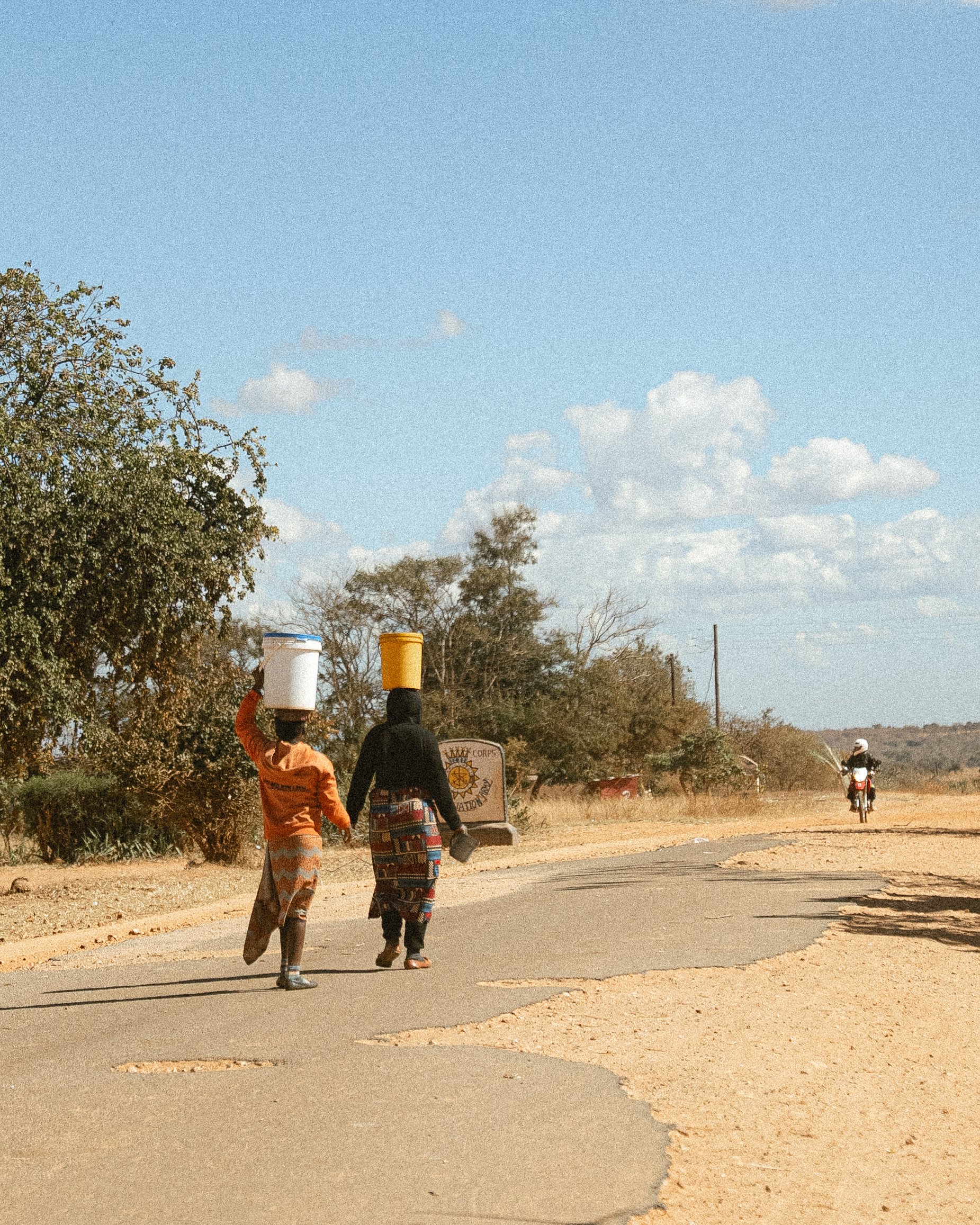 Two women walking on a rural road, balancing buckets on their heads, in a dry, semi-arid landscape with scattered trees and a blue sky with clouds.