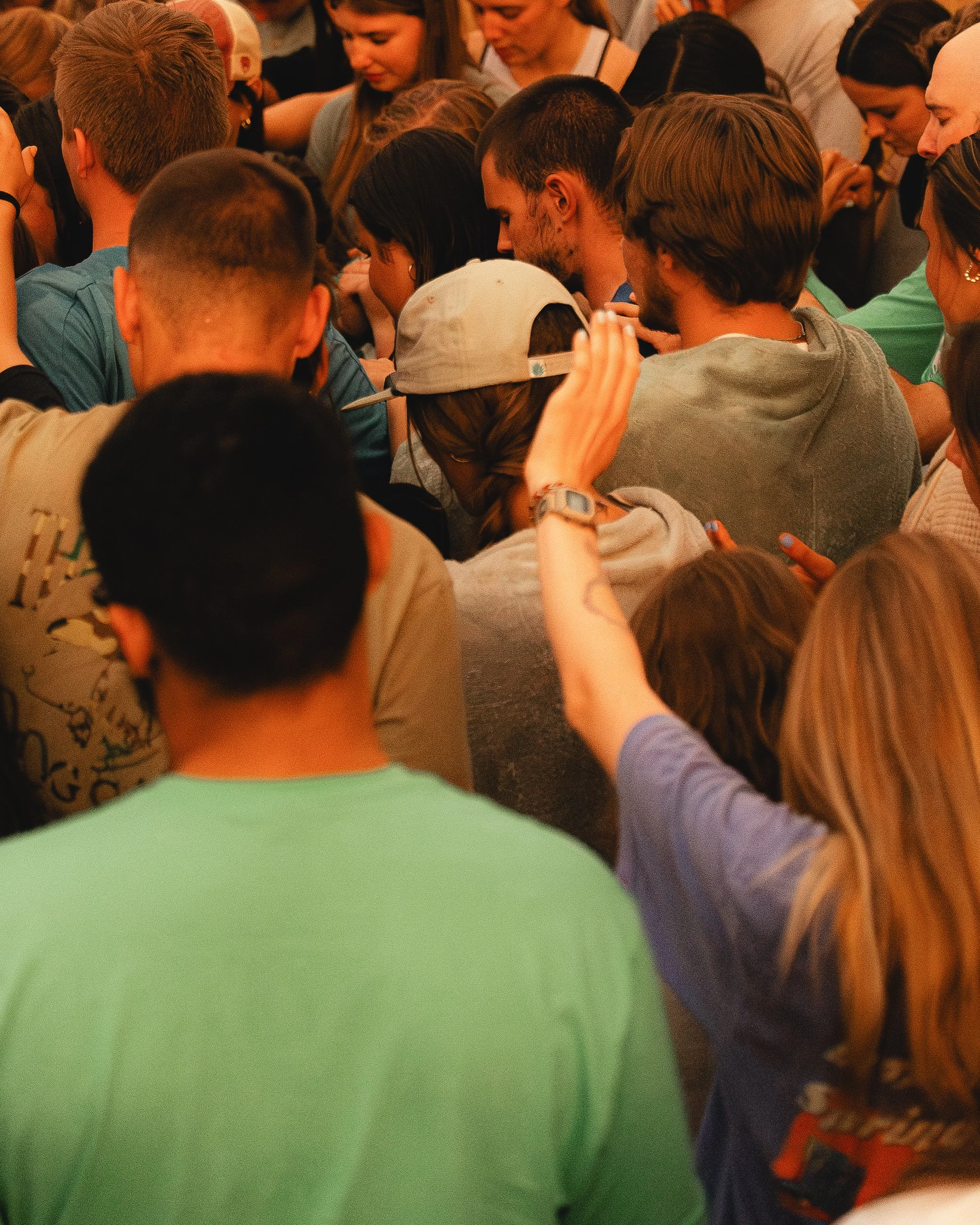 Crowd of young people closely gathered, many with bowed heads, some with hands raised or touching their heads, in a communal, prayerful or contemplative state.