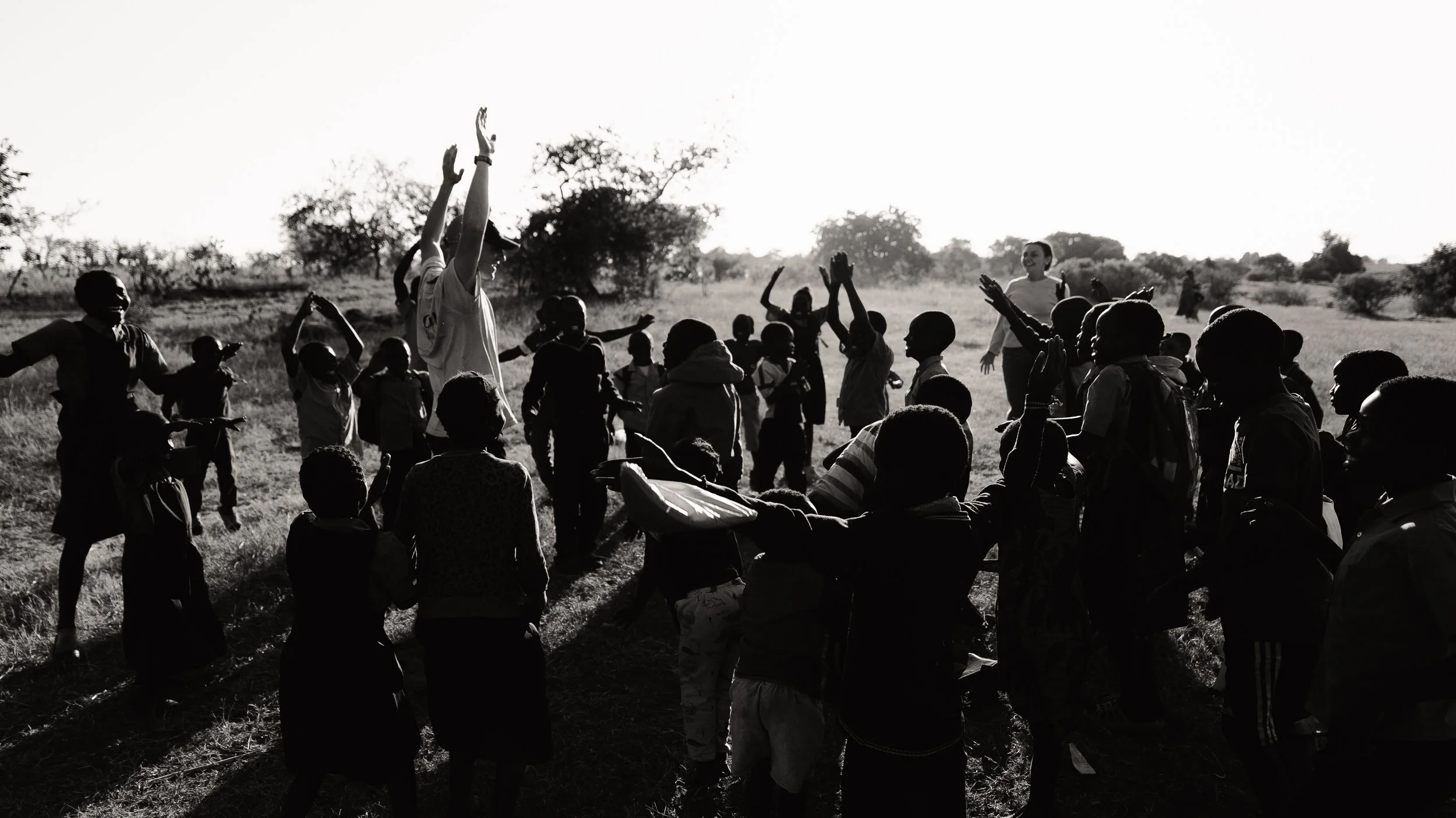 A group of children and a few adults outdoors in a grassy field, some raising their hands and others dancing or moving around, with the sun setting or rising in the background.