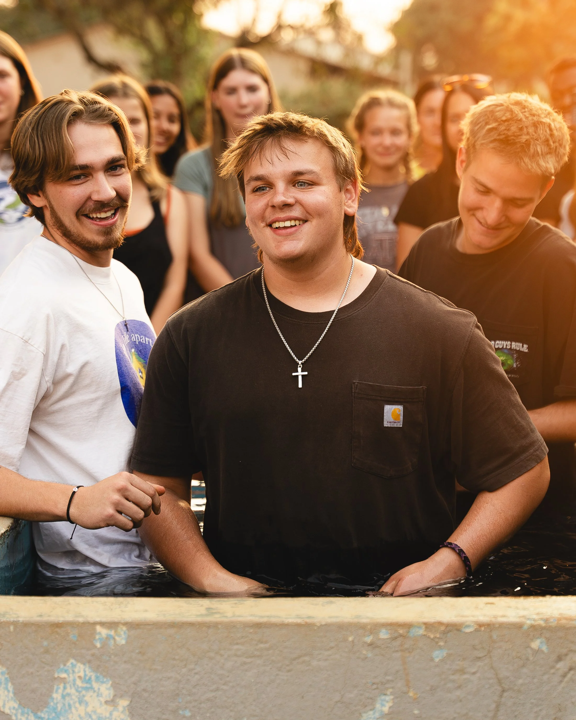 Group of young people gathered outdoors, with three young men in the foreground, one wearing a cross necklace, standing in water and smiling, as the sun sets in the background.