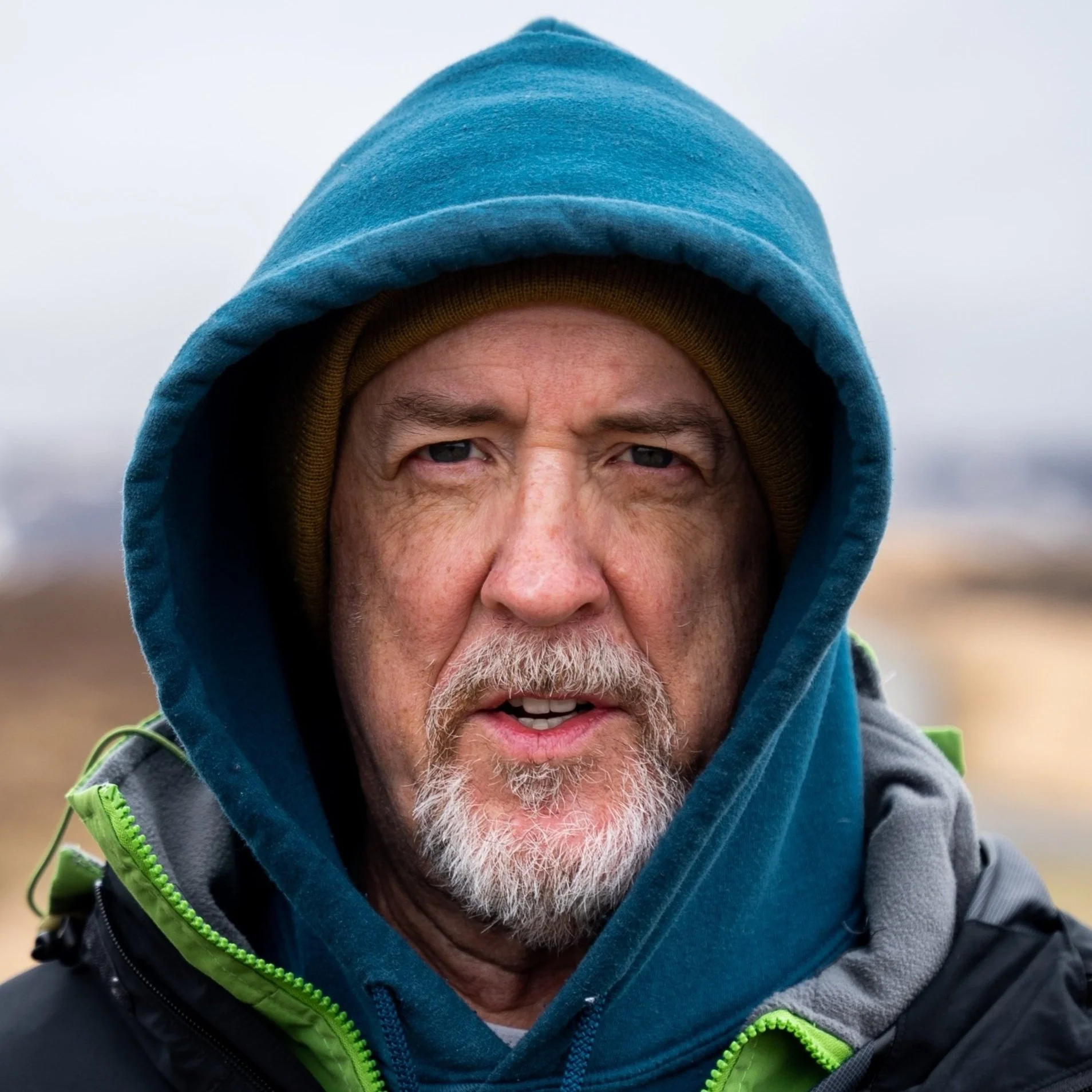 Close-up of a middle-aged man with a gray beard, wearing a blue hoodie and a brown beanie, outdoors on a cloudy day.
