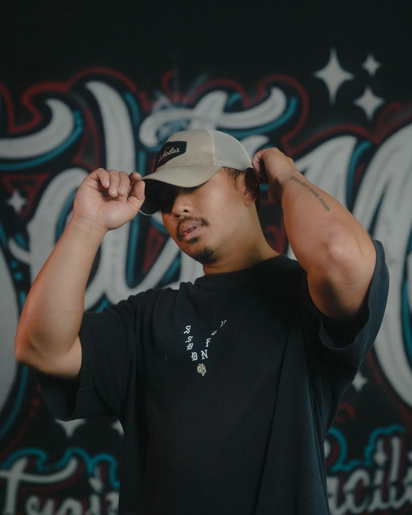A man wearing a beige cap, black t-shirt, and a tattoo on his left arm is adjusting the cap with his right hand while standing in front of a colorful graffiti wall.