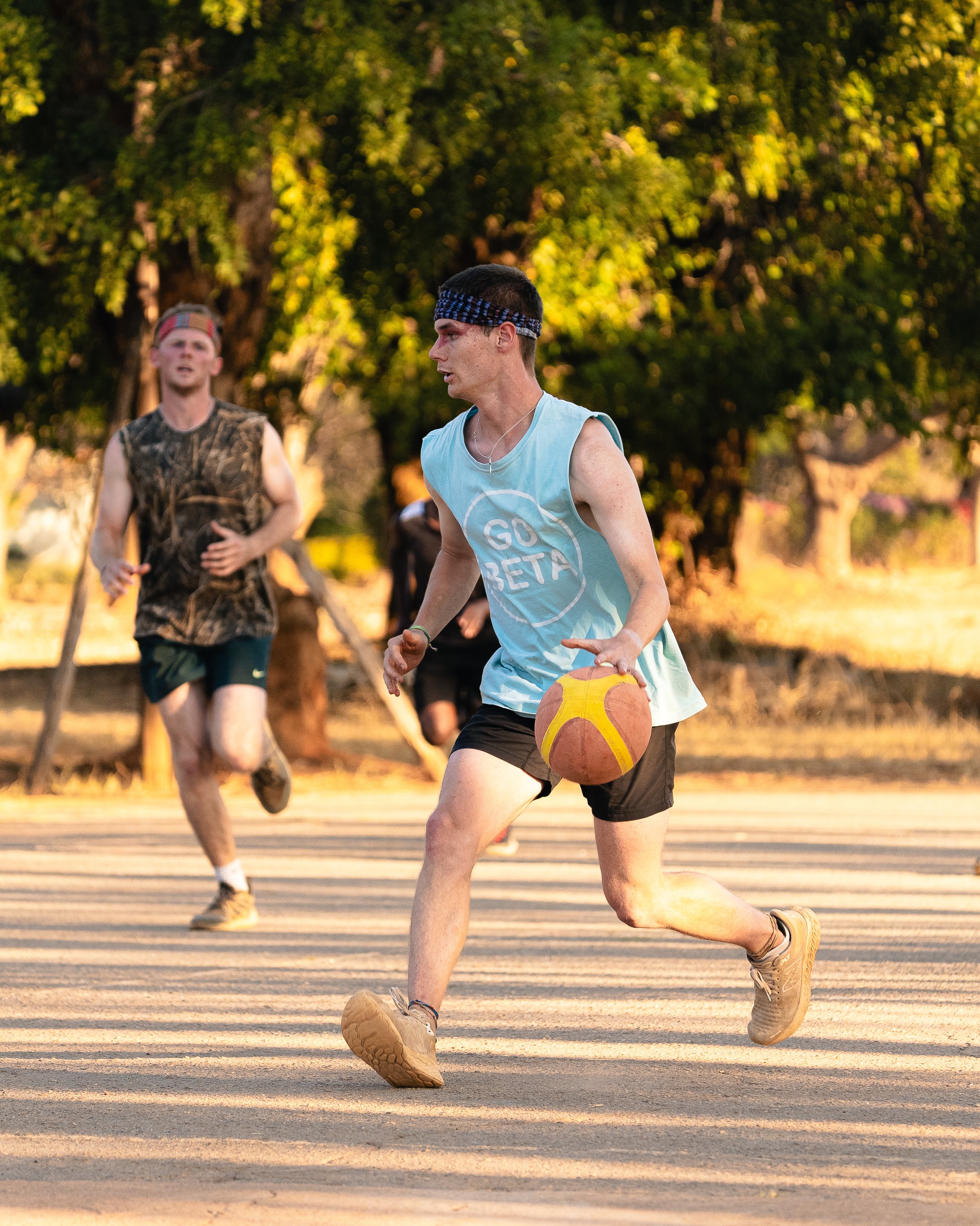 Young man playing basketball outdoors during sunset, wearing a blue tank top with 'GO BETA' and a bandana, with others running in the background.