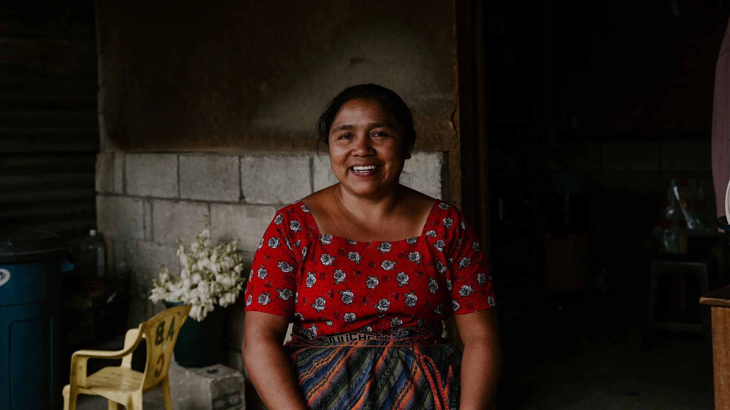 Smiling woman in red floral top sitting indoors, with a bouquet of white flowers in the background.