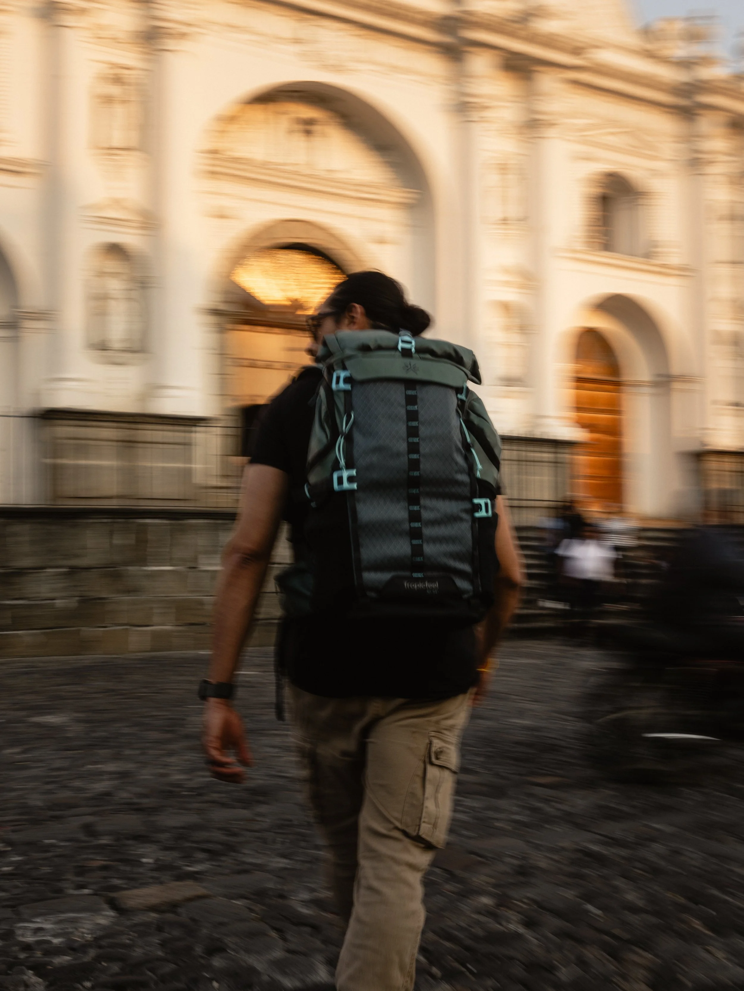 Person wearing a black shirt and beige cargo pants carrying a large teal backpack walking on cobblestone street in front of an ornate white building.