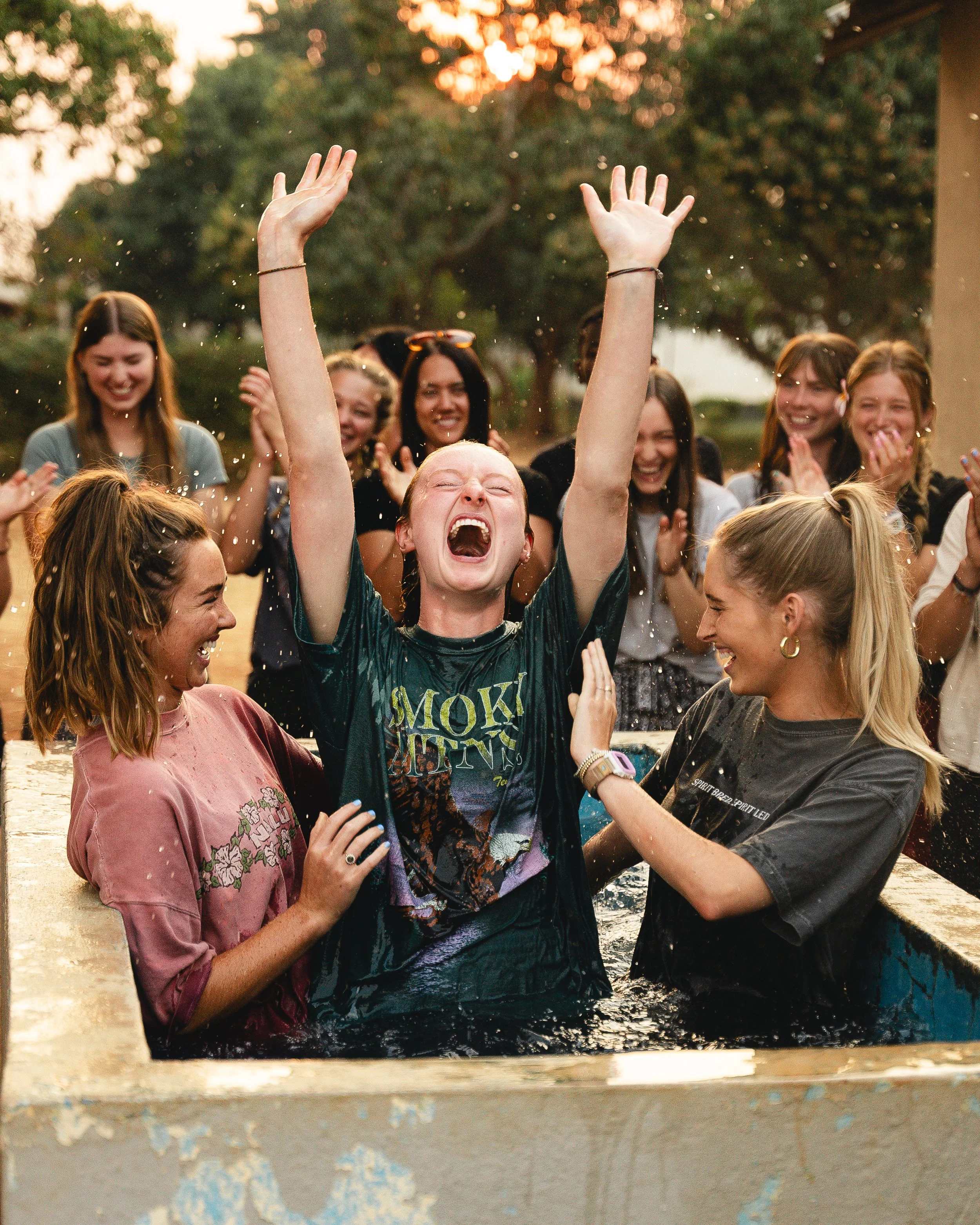 Group of young women celebrating outdoors during sunset, with one woman in a baptismal pool raising her arms in joy while others cheer around her