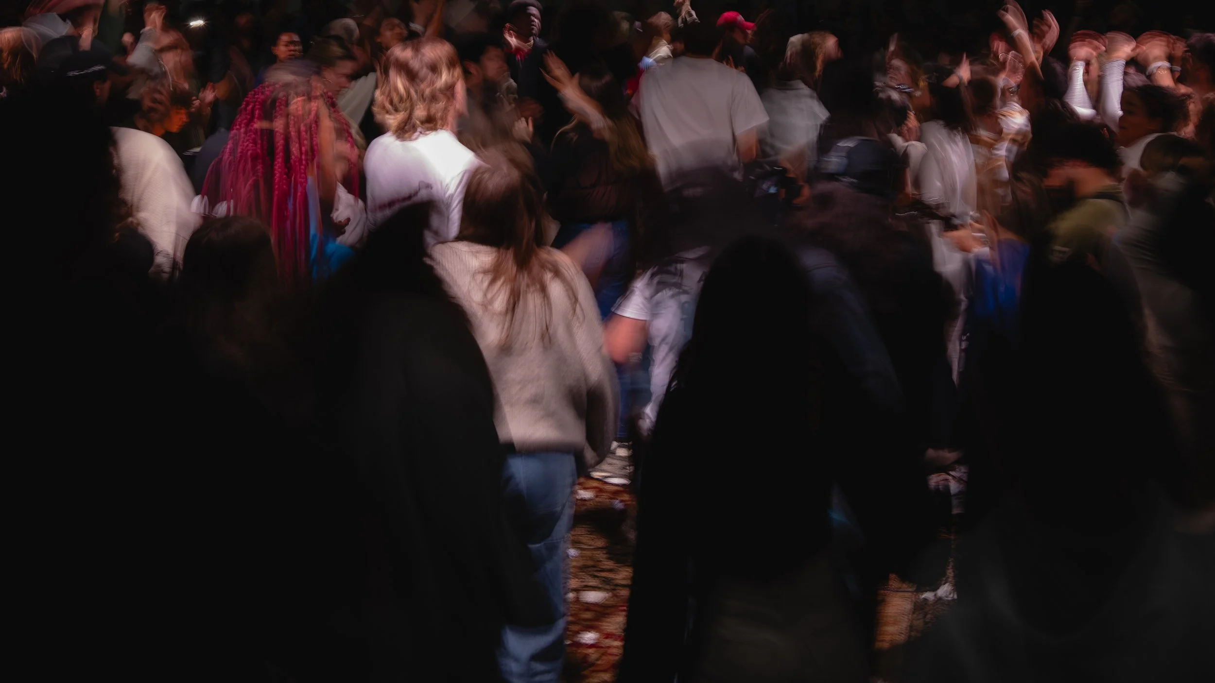 Crowd of people gathered closely together at an indoor event, some with raised hands and colorful hair, blurry and dark in the background.