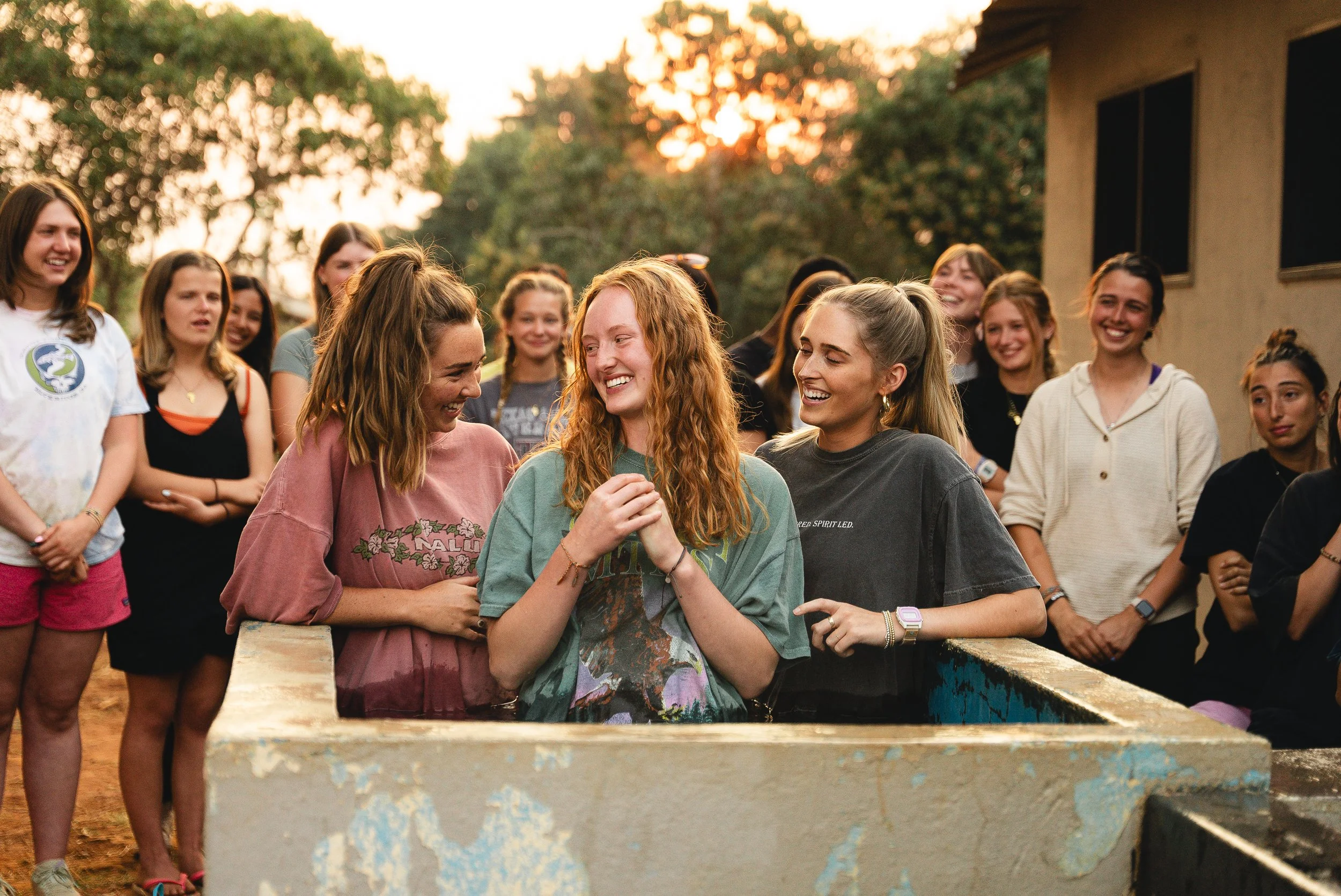 A group of young women laughing and talking outdoors at sunset, around a small pool or trough, with trees and a building in the background.