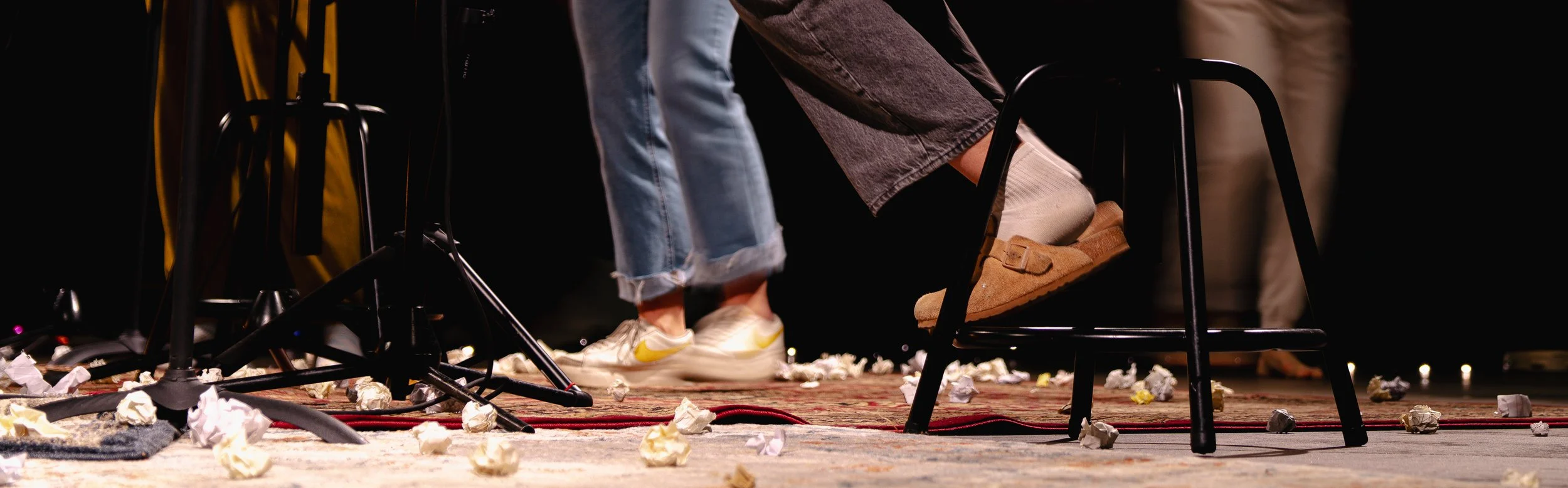 People walking on a stage with crumpled paper scattered on the floor, surrounded by chairs and equipment.