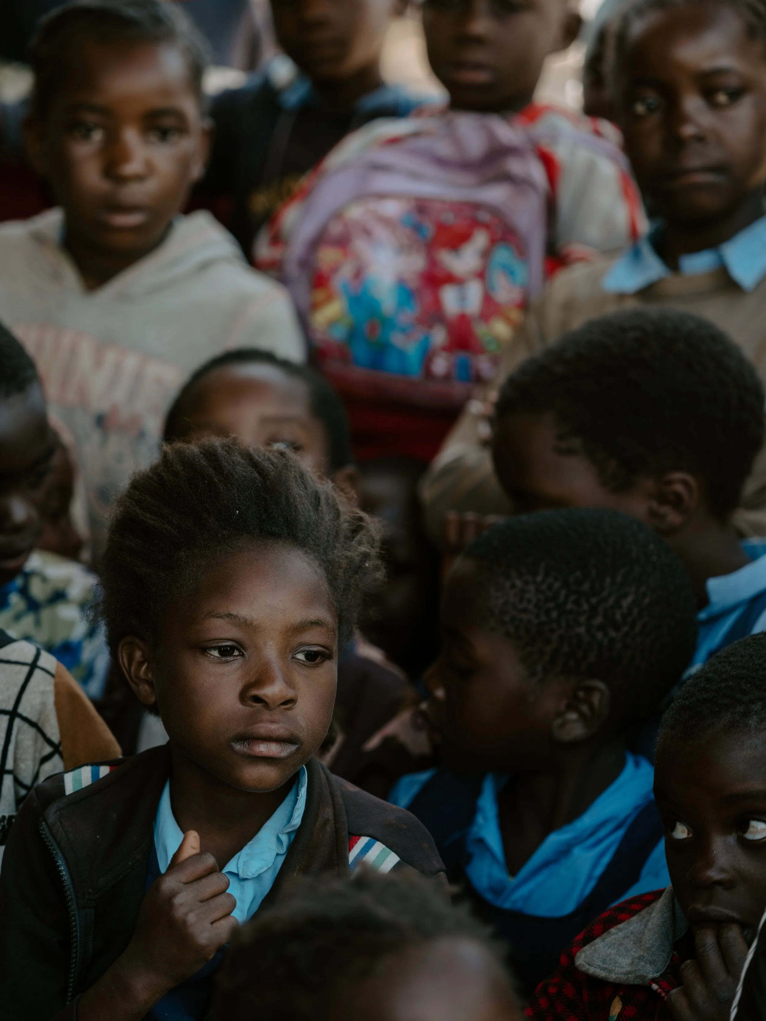 A group of young children, mostly African, gathered closely together, some with serious or contemplative expressions, in a crowded setting.