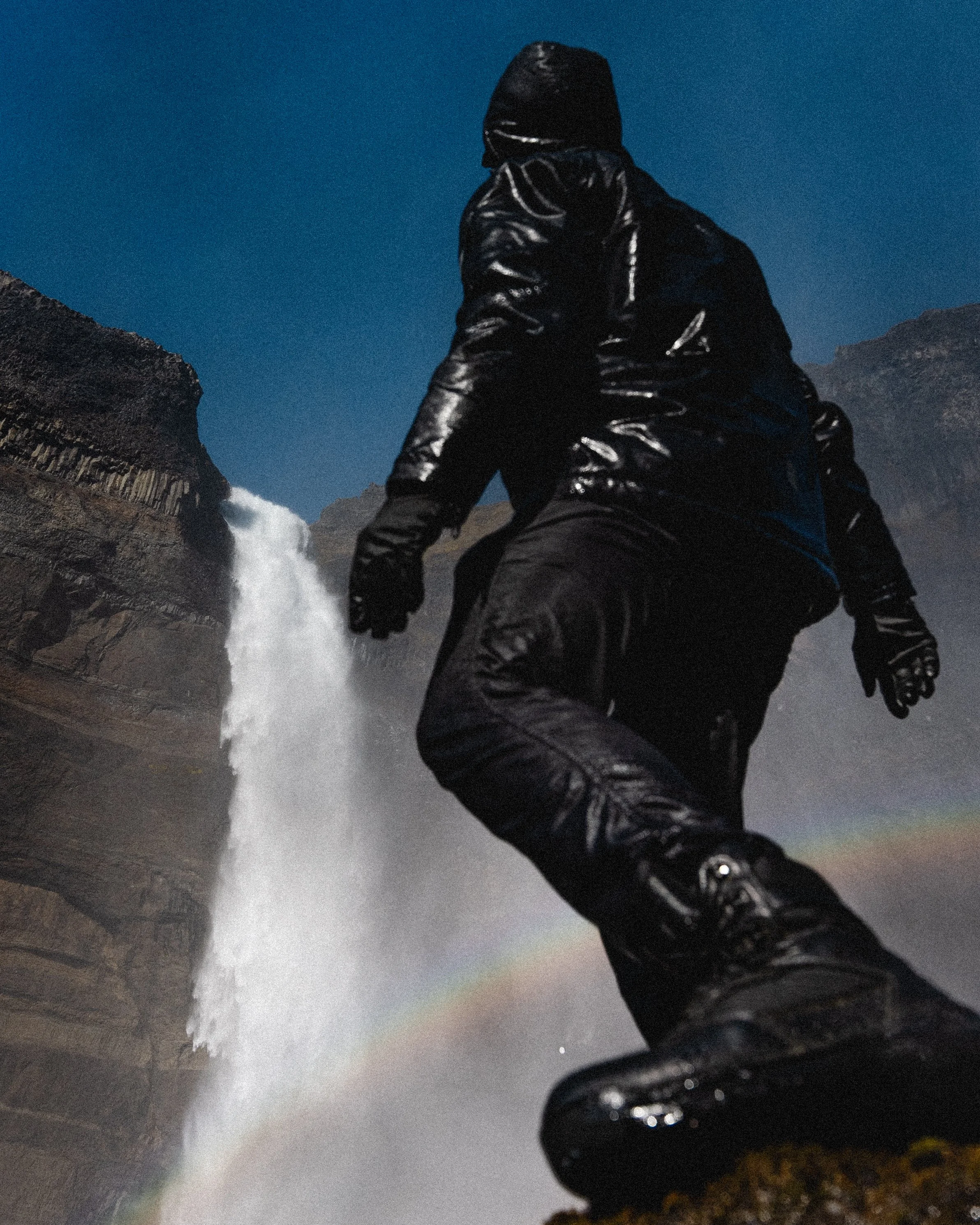 A person wearing black waterproof clothing sitting near a waterfall with a visible rainbow in the mist.