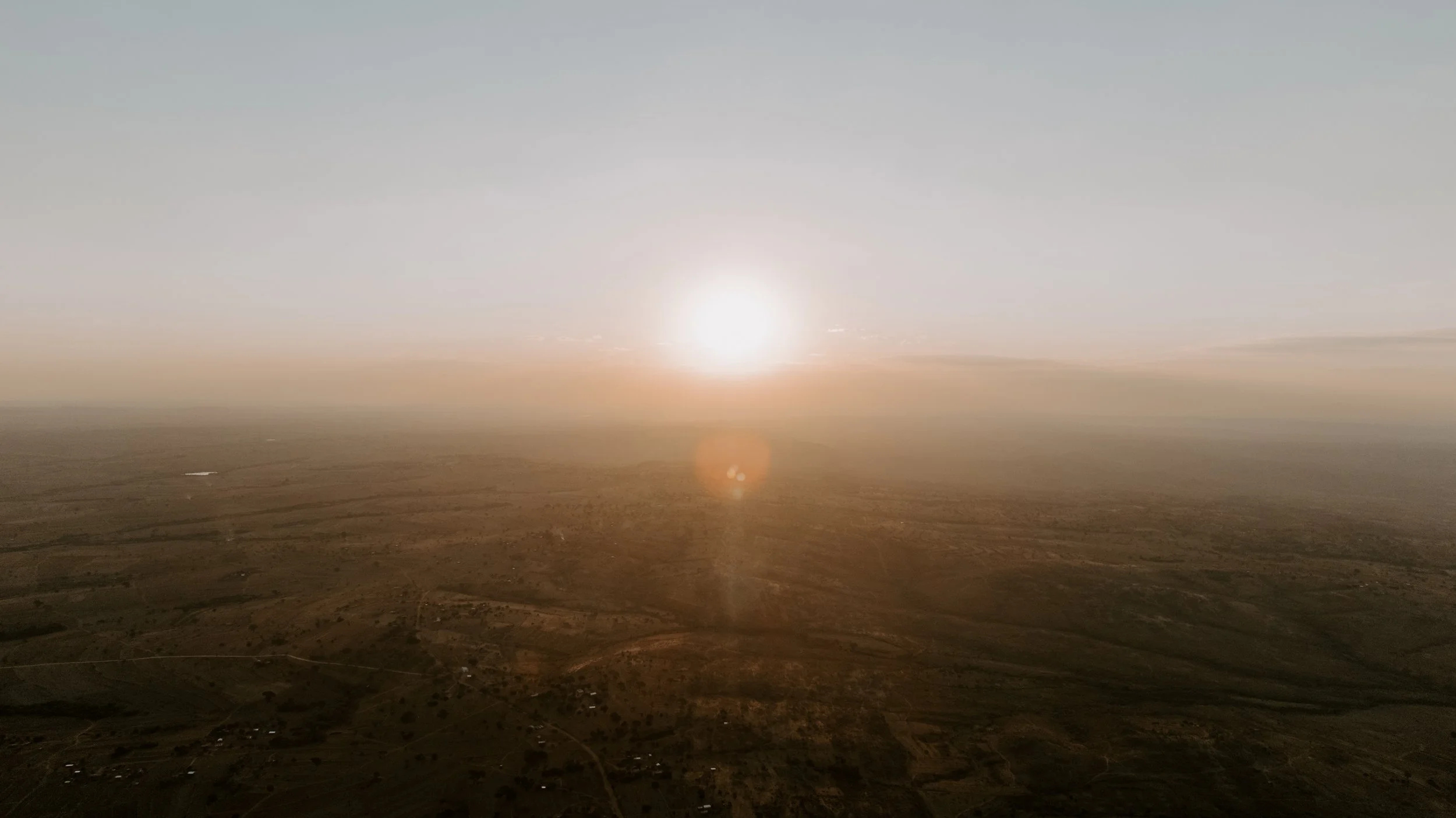 A vast, flat, and arid landscape with a setting or rising sun near the horizon, creating a warm glow and lens flare in the sky.