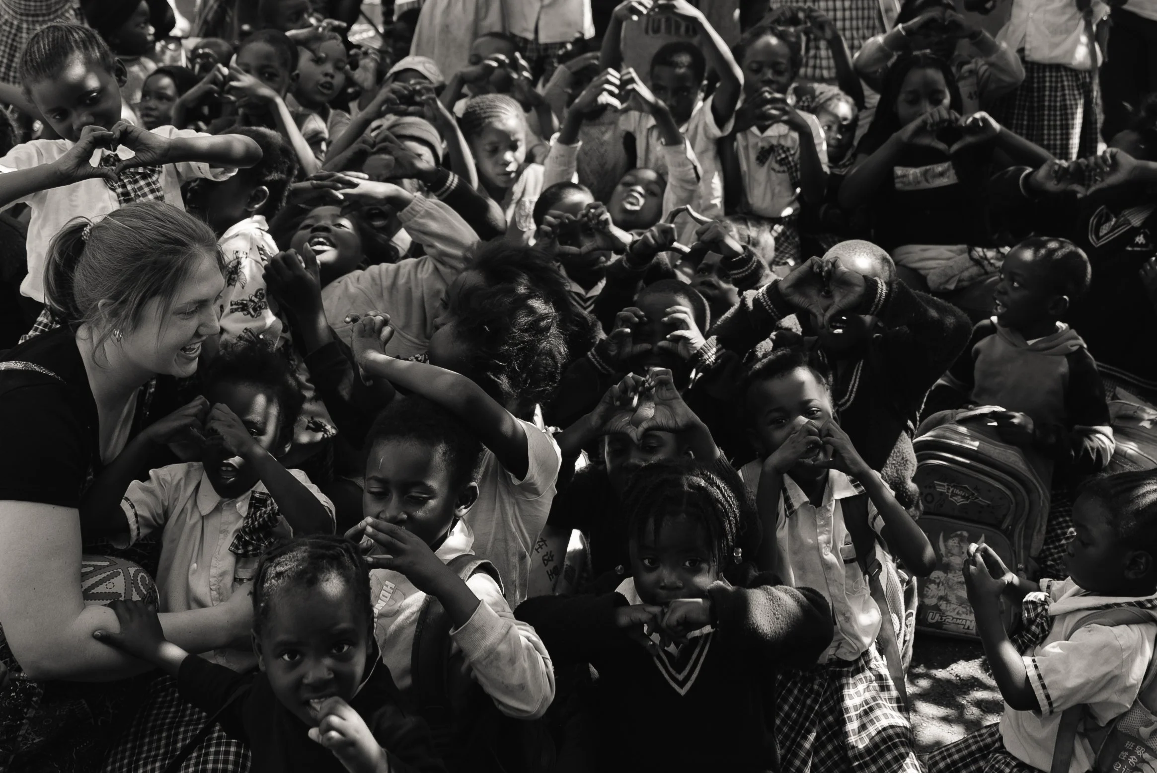 A large group of children and a woman engaging in a fun activity outdoors, with many making heart shapes with their hands.