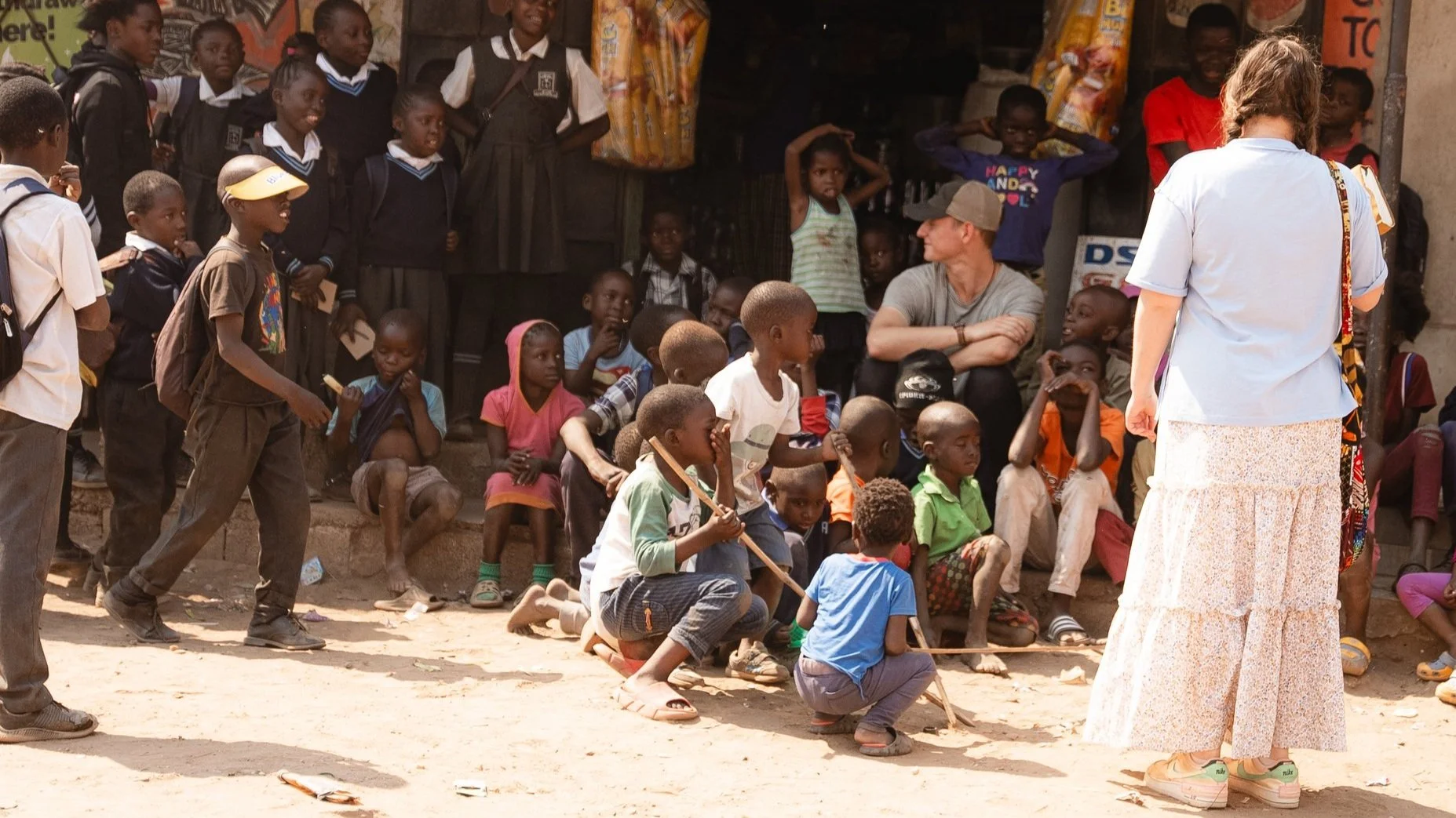 A group of children sitting and standing outdoors, with a woman in a light blue shirt and long skirt facing them, and a man in a gray shirt and cap sitting on the steps. The children are watching attentively while some are squatting and holding stick