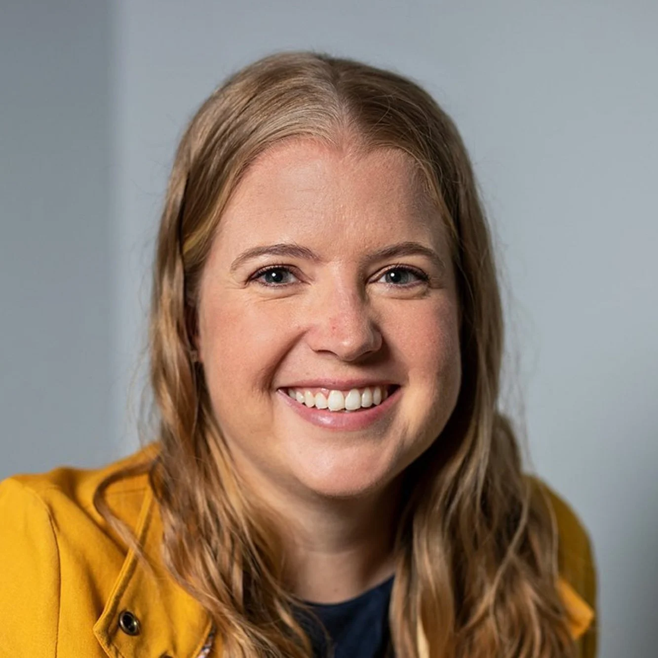 Close-up portrait of a smiling woman with wavy light brown hair, wearing a mustard yellow jacket over a dark shirt, against a neutral background.