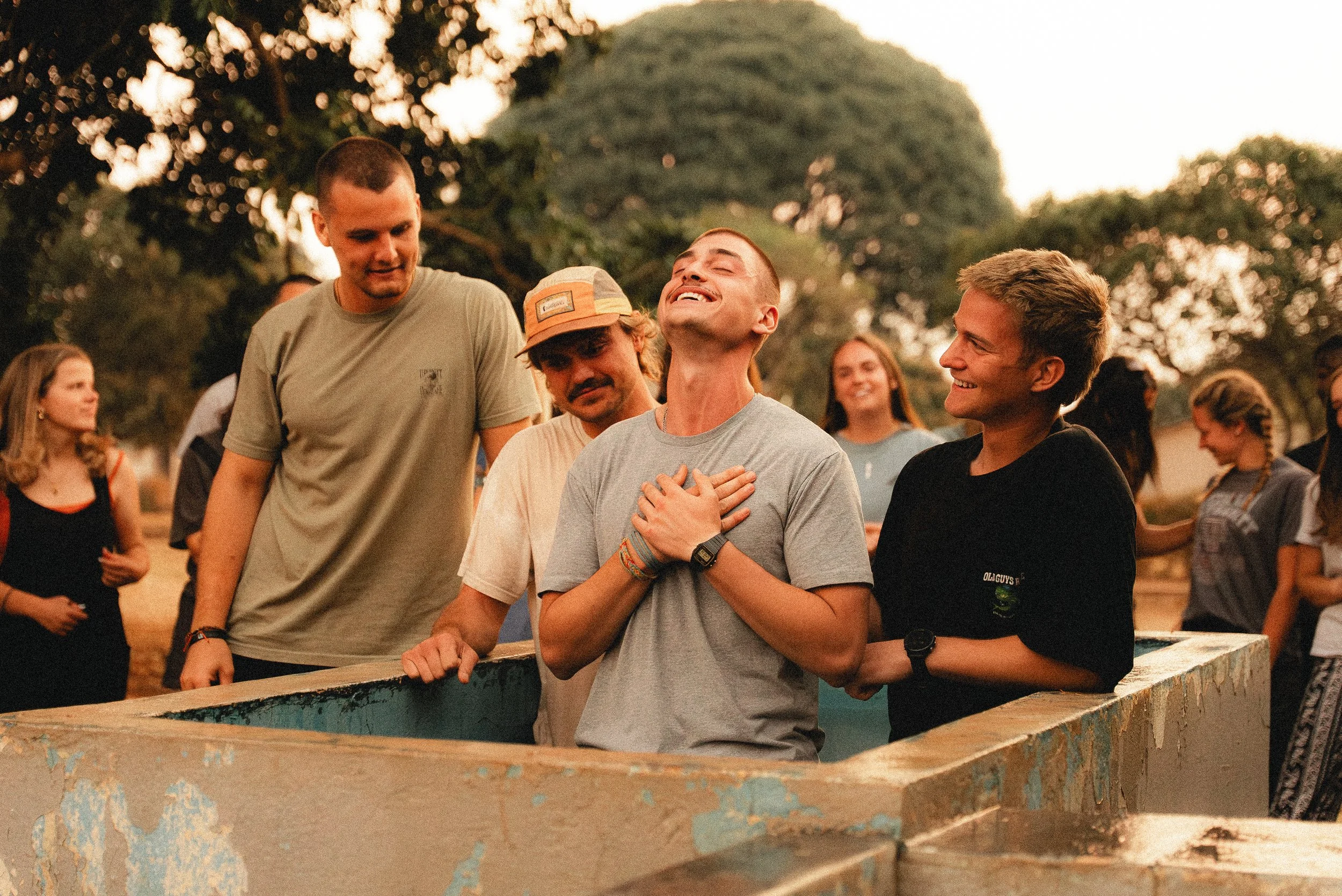A group of young people outdoors, with some around a concrete structure, smiling and enjoying themselves during sunset.