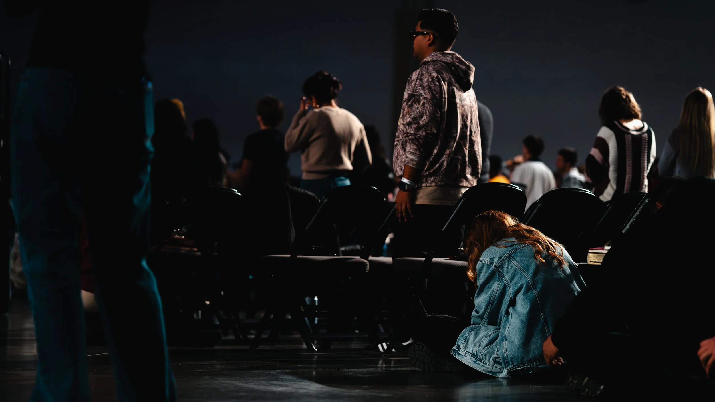 Young girl with red hair wearing a denim jacket sitting on the floor, head bowed, next to empty chairs in a dark, crowded room.