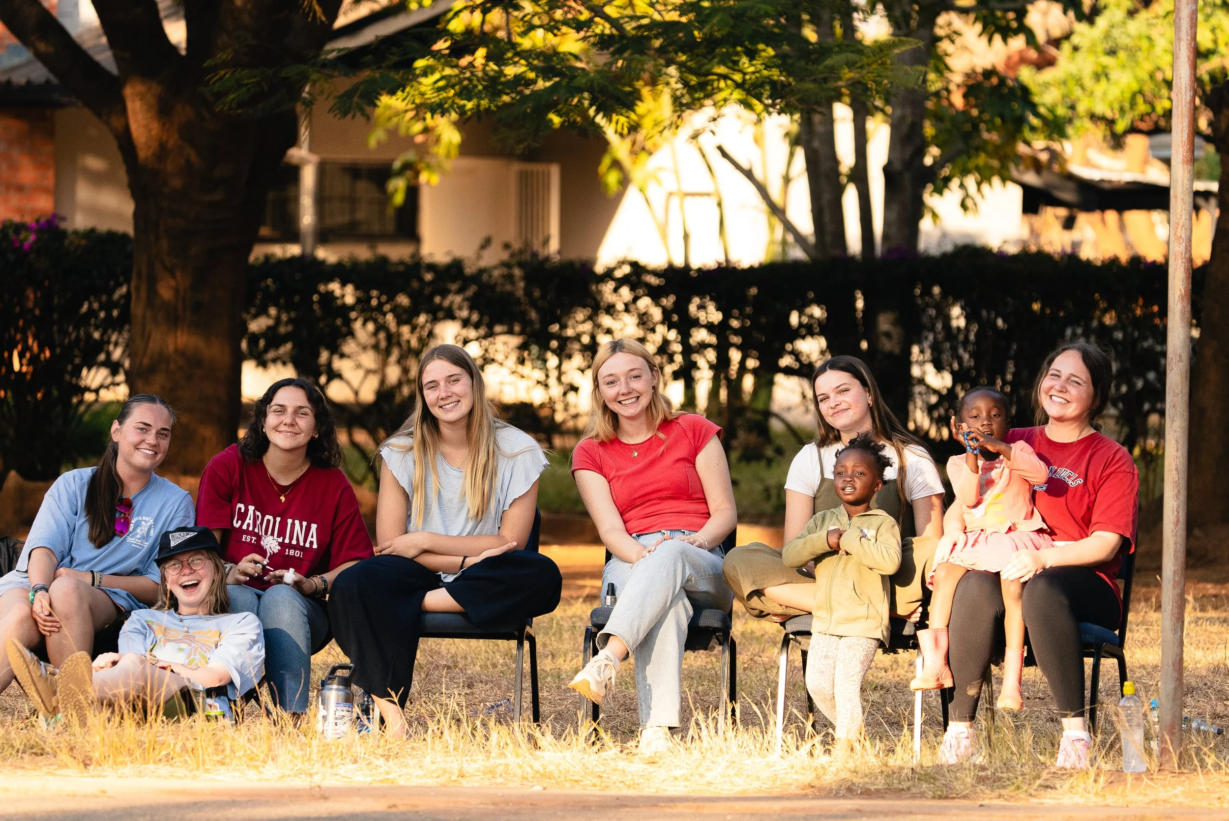 A group of nine smiling women and children sitting outdoors during sunset, with trees and a hedge in the background.
