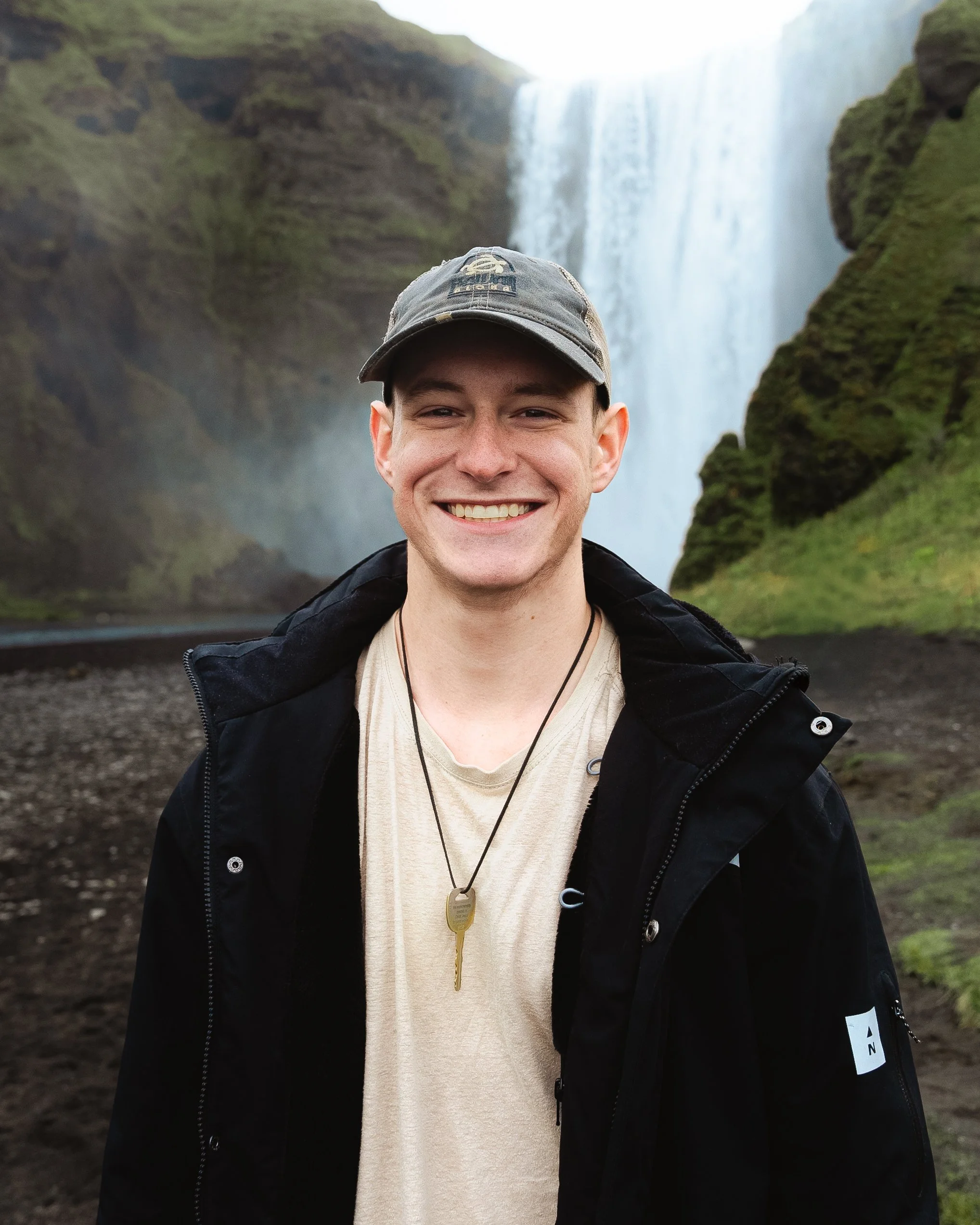 A young man smiling in front of a large waterfall with green moss-covered cliffs.