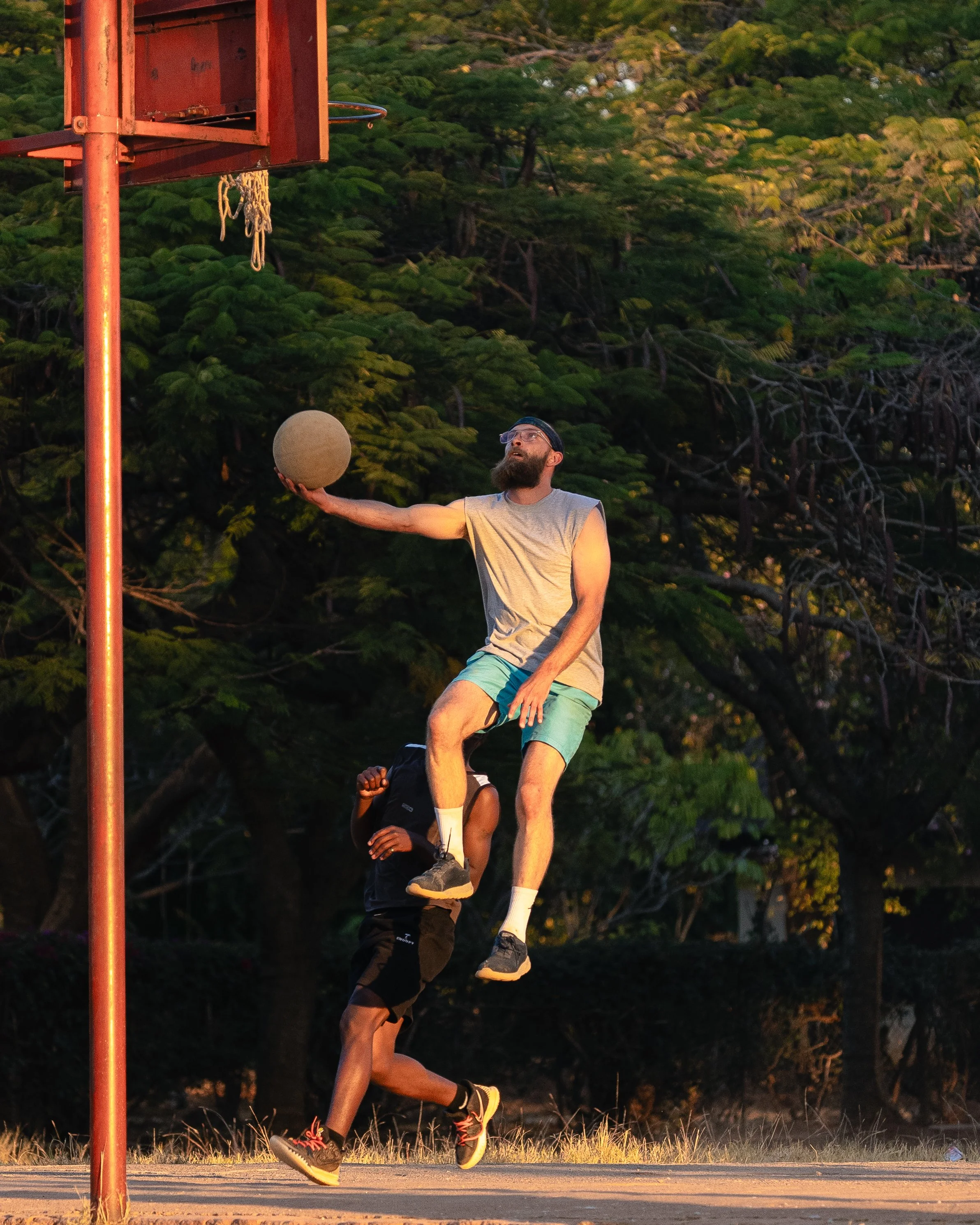 A man jumping to block a basketball shot near the hoop on an outdoor court, with trees in the background.