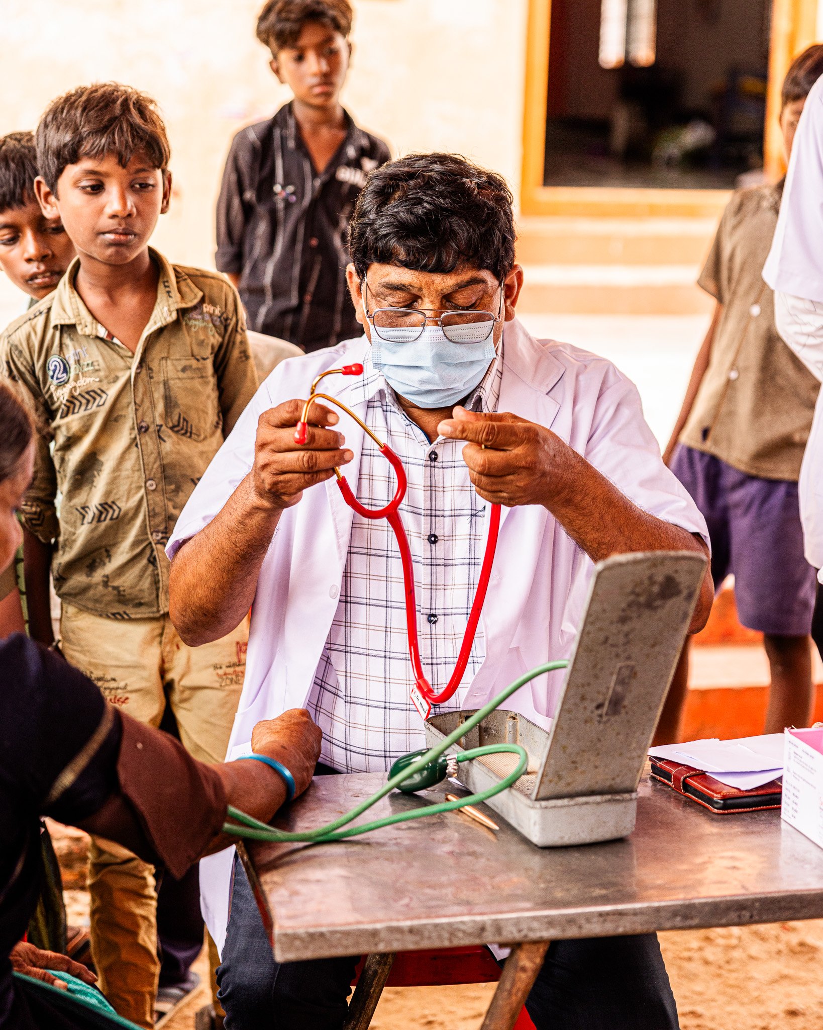 A doctor wearing a face mask and glasses demonstrates using a stethoscope to a patient in an outdoor setting, with children watching.