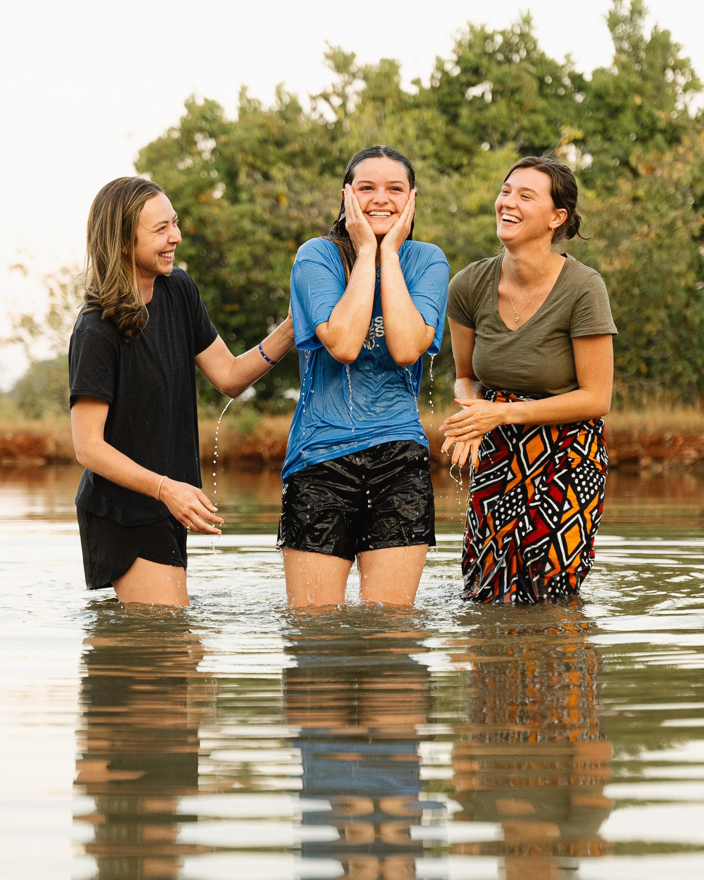 Three women standing in a body of water, smiling and laughing together, with trees in the background.