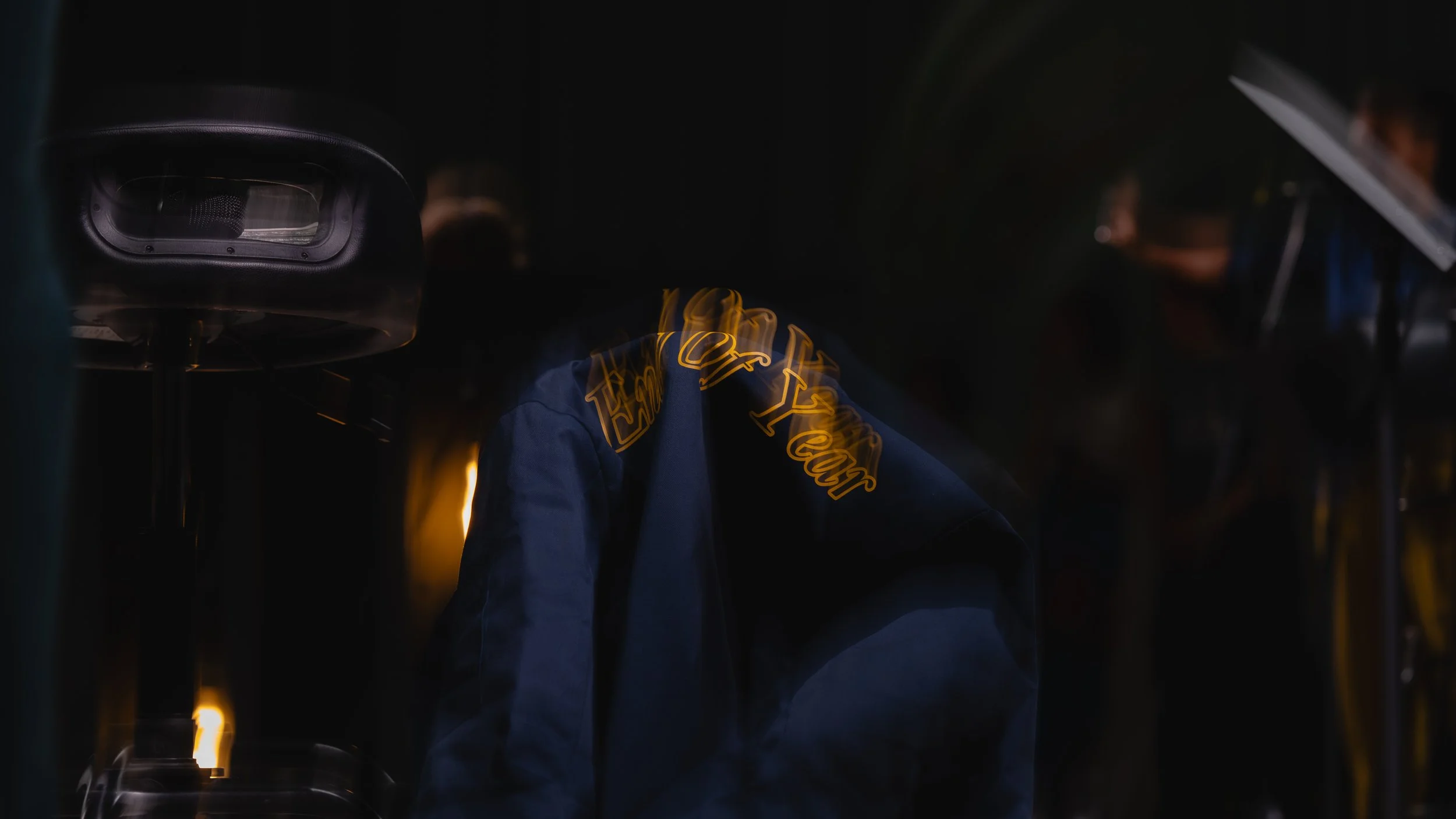 A dark scene with a person wearing a blue jacket that has yellow lettering, surrounded by dance floor lighting and equipment, possibly at a DJ booth or stage.