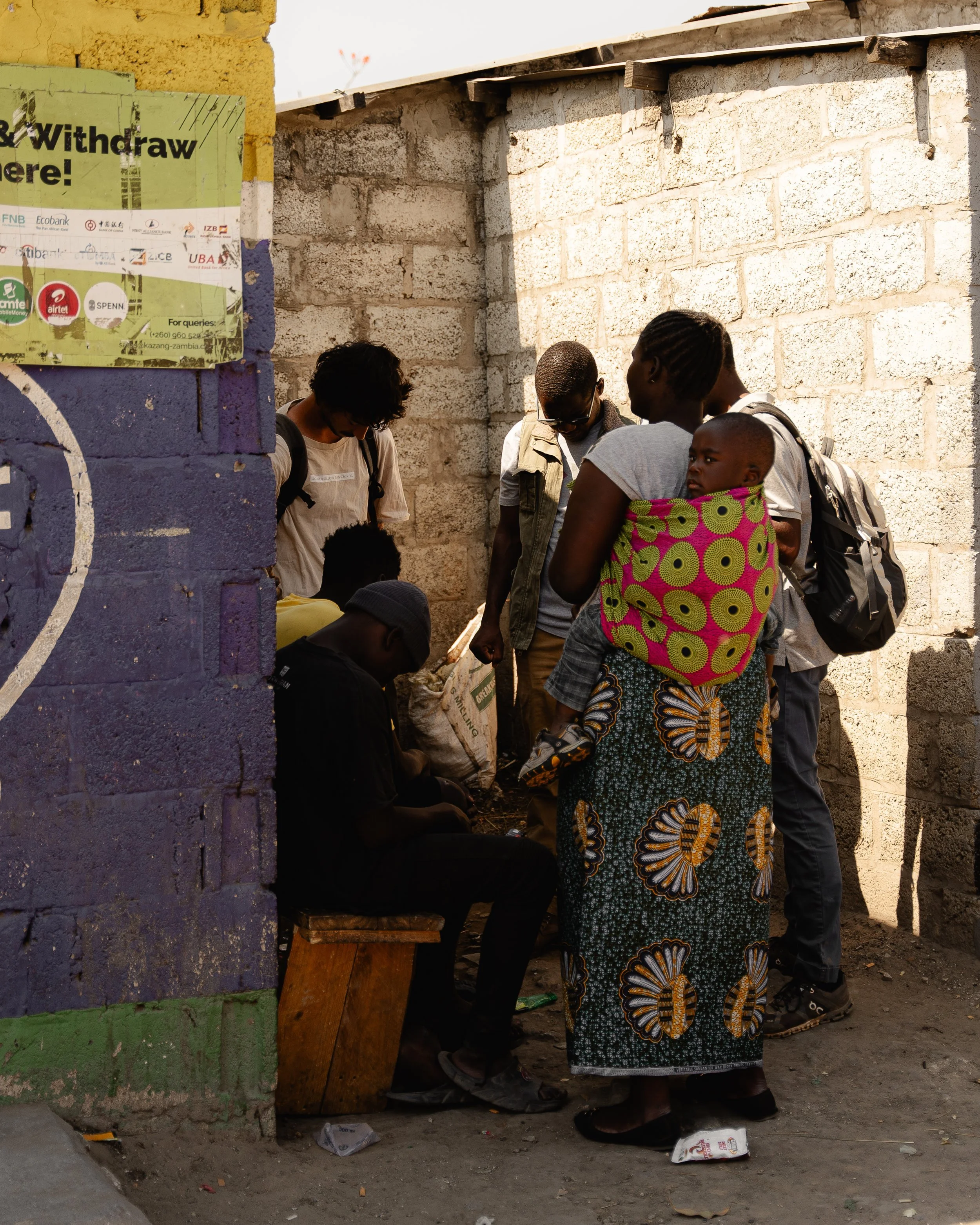 A group of people gathered in an outdoor corner, with some sitting on a bench and others standing, engaged in conversation or an activity, next to a brick wall with a purple and green painted section and a cardboard sign partially visible.
