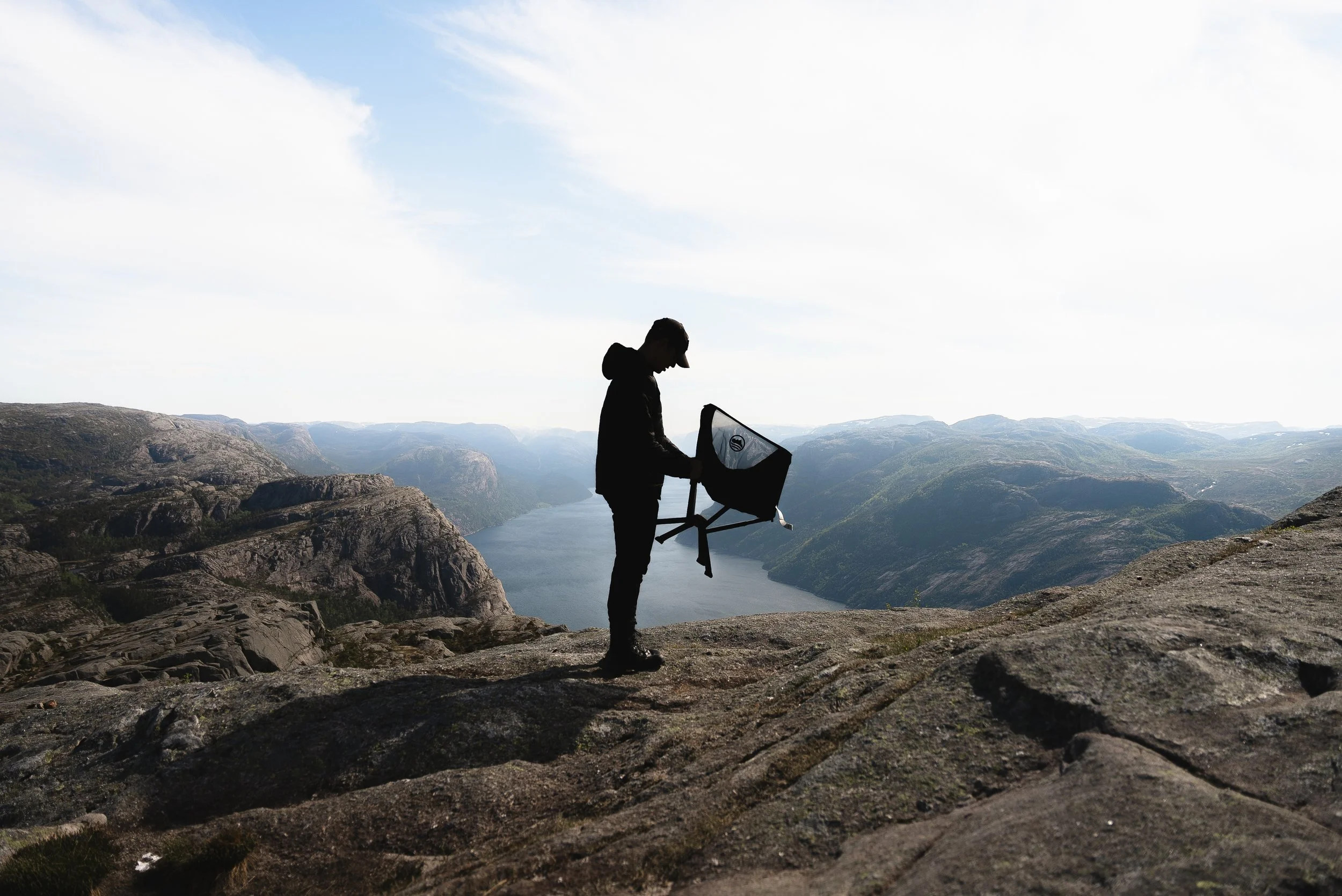 Silhouette of a person standing on rocky terrain, looking down at a baby stroller, with a backdrop of mountains and a cloudy sky.