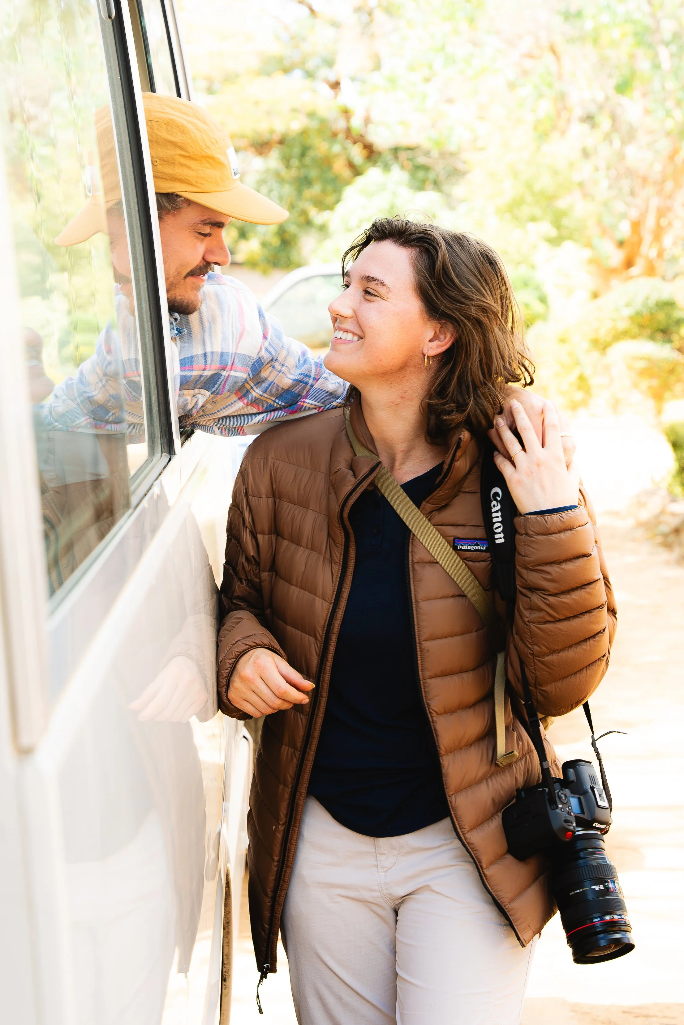 A woman with a camera around her neck smiling and touching her hair, standing outside near a woman leaning out of a vehicle window, sharing a moment of laughter.