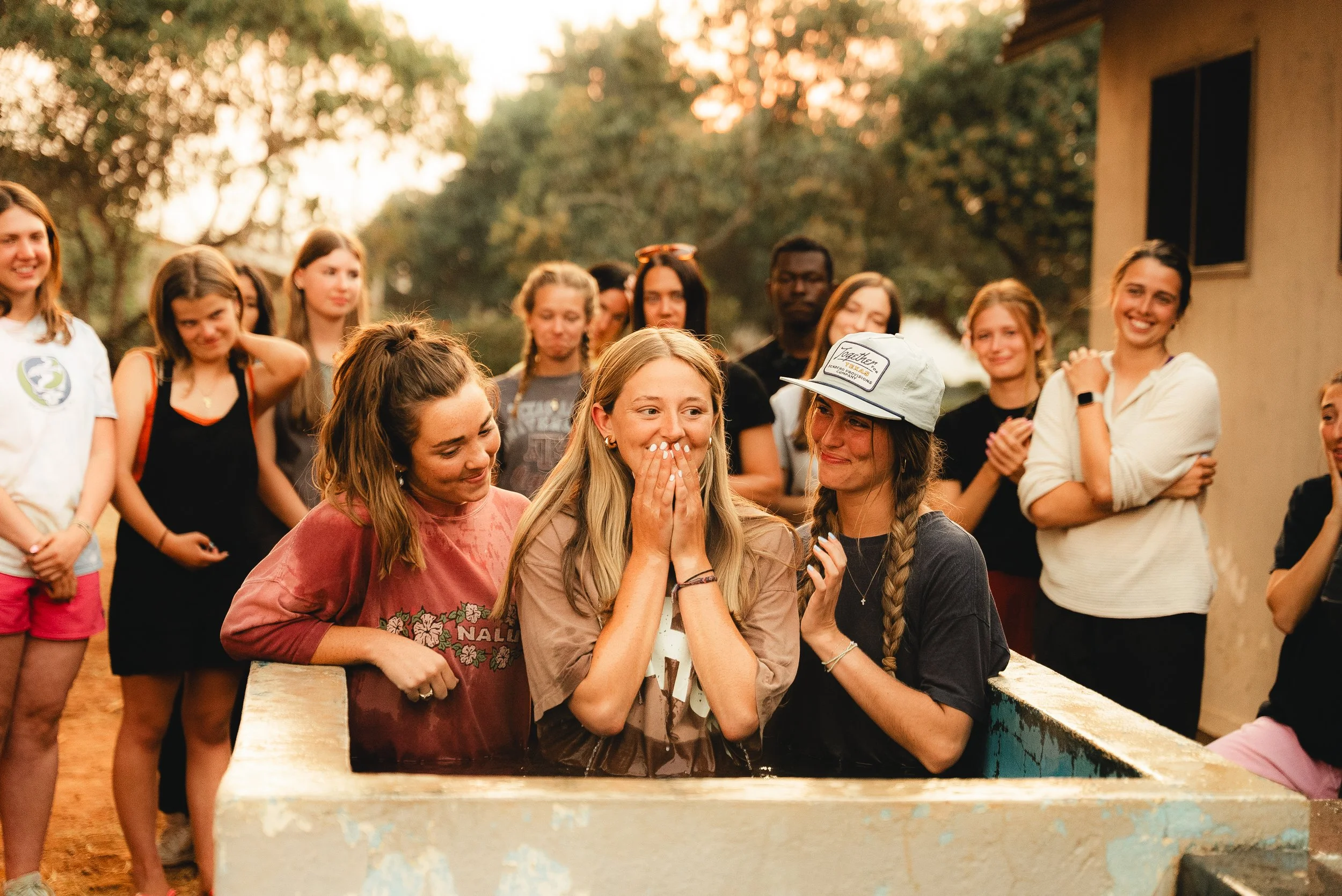 A group of young people gathered outdoors around a girl who is emerging from a water tank during a baptism ceremony, with others smiling and watching
