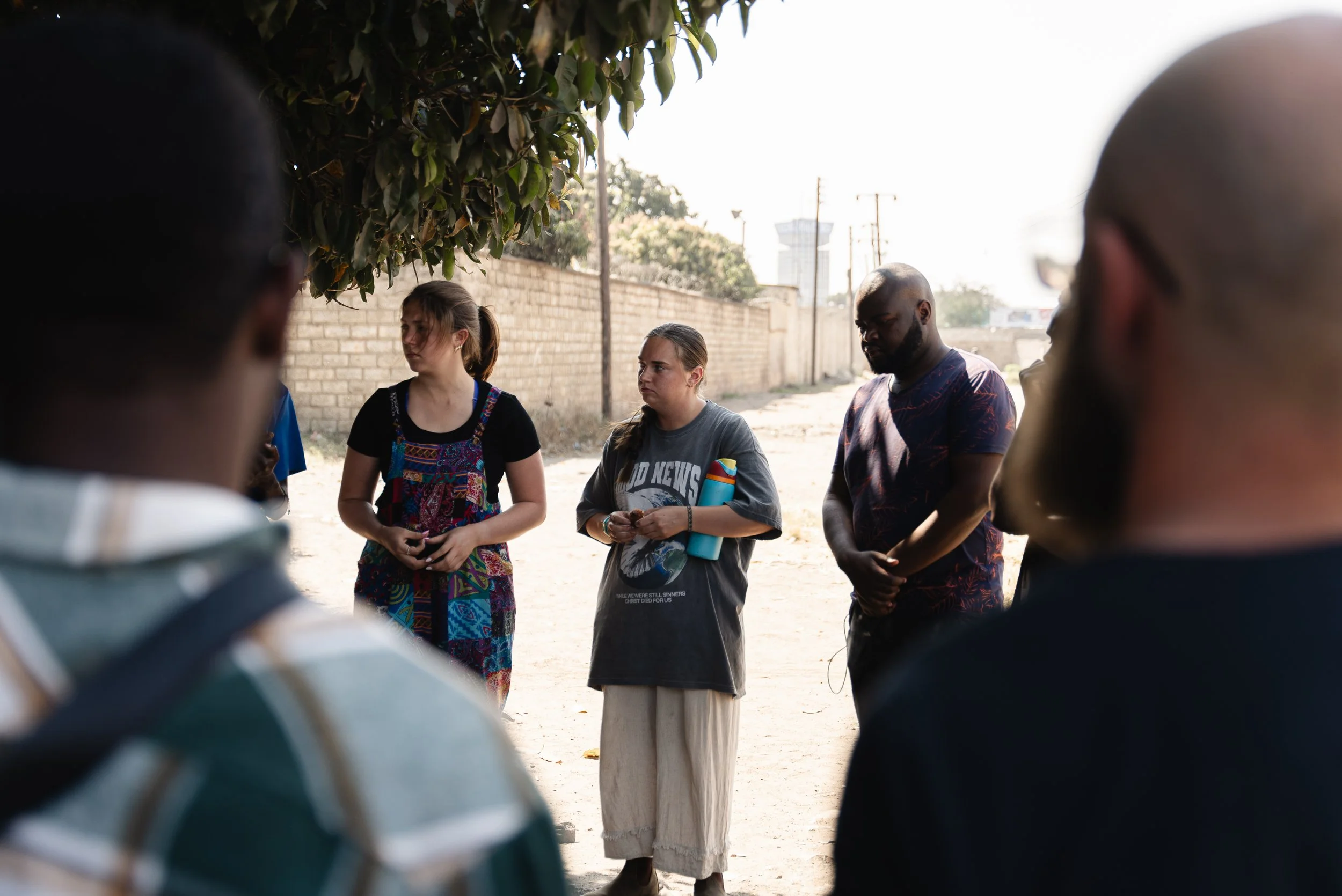 Group of people standing outdoors in a circle, listening to a woman speaking, with some trees and a brick wall in the background.